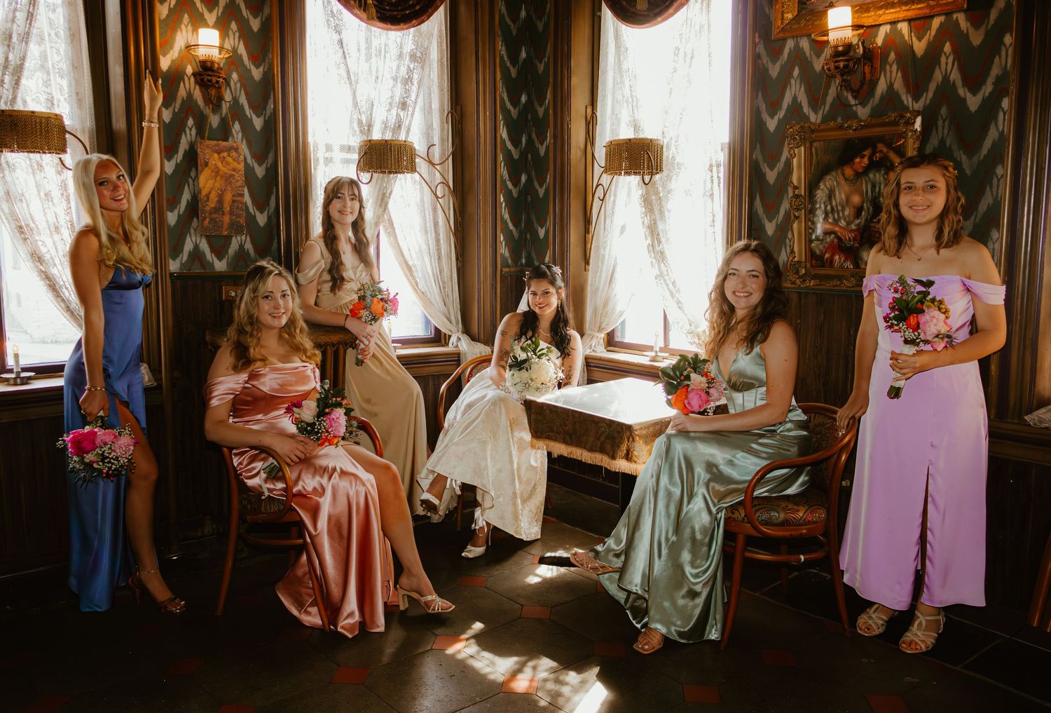 Five bridesmaids in colorful dresses pose with bouquets in a vintage-styled room with ornate decor.