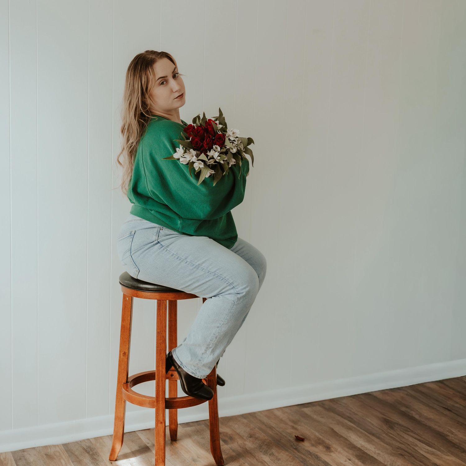 Woman in green sweatshirt and light jeans sits on wooden stool holding red and white bouquet against white wall.