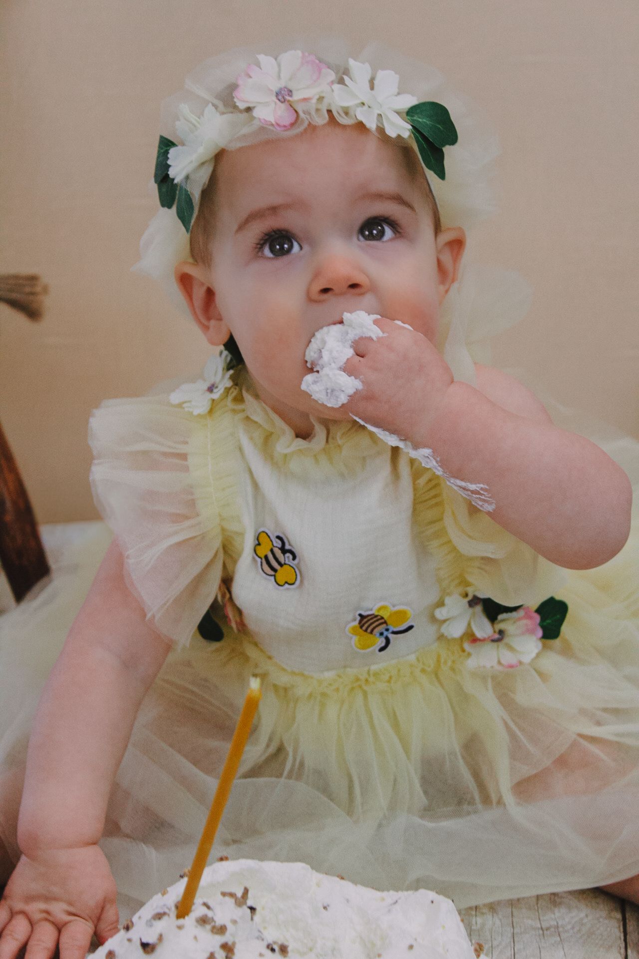 A baby in yellow dress and flower crown enjoys cake during a celebration.