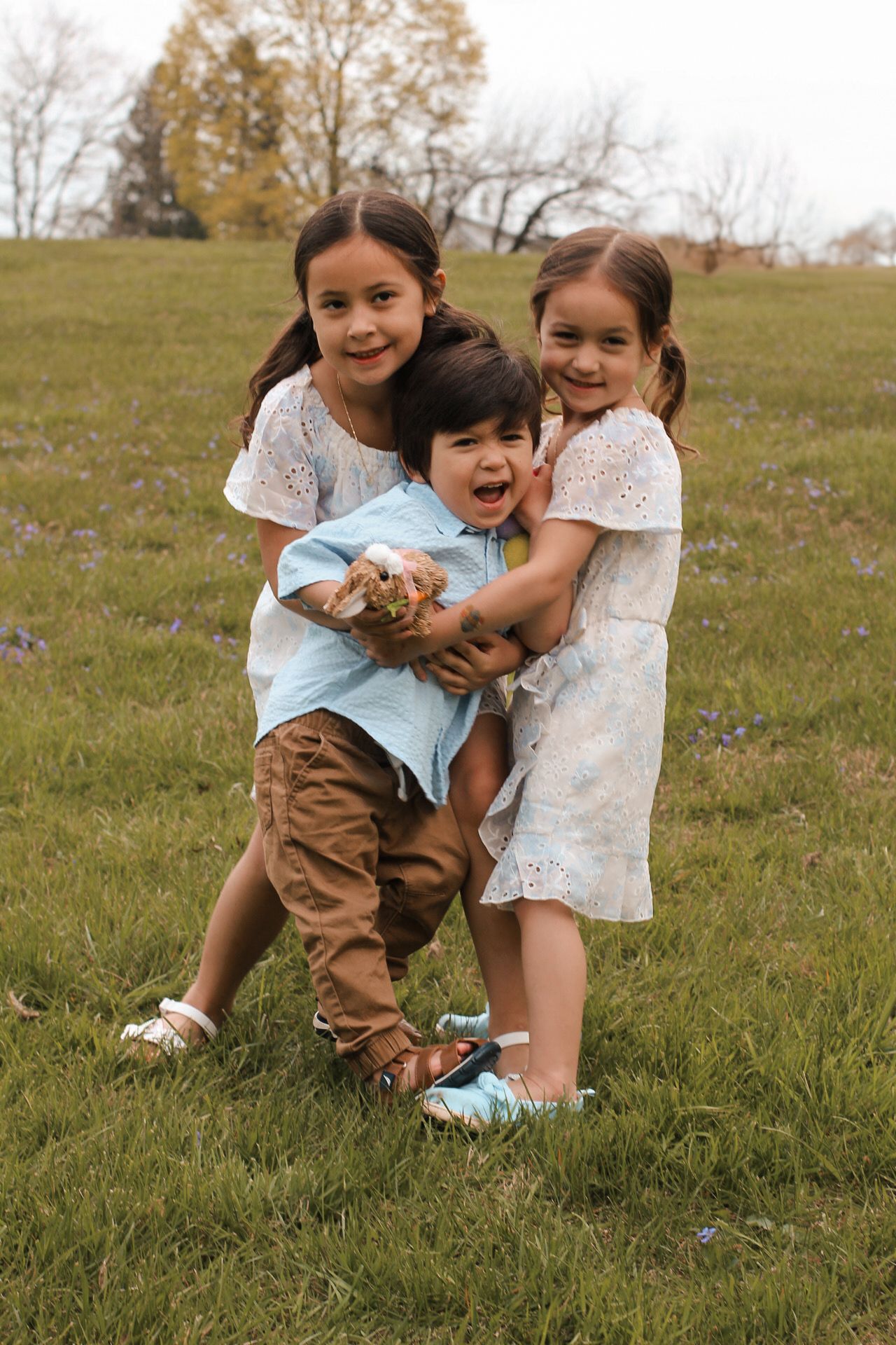 Three siblings in casual attire play together on a grassy hill during autumn while sharing a teddy bear.