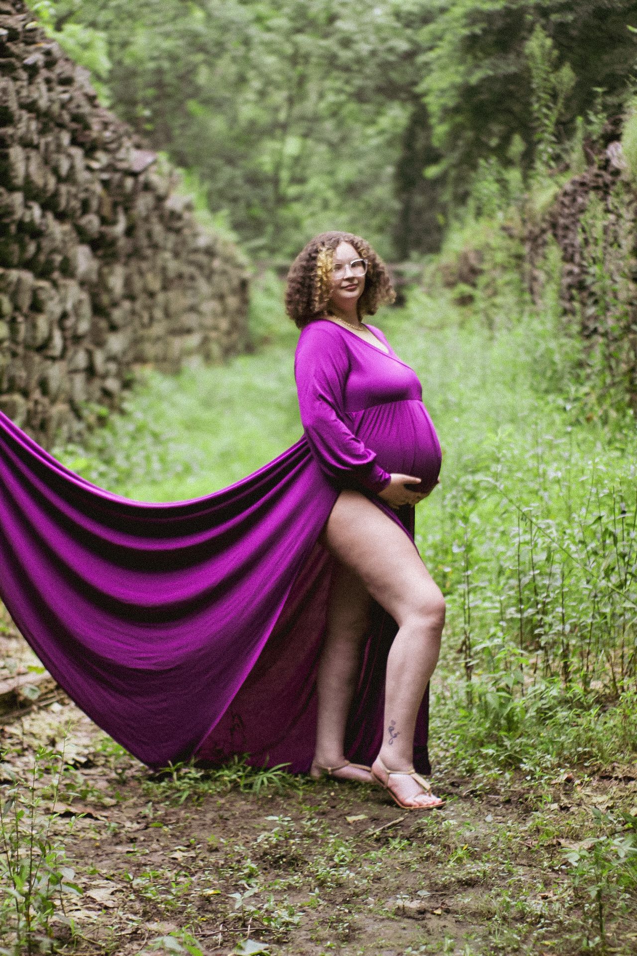 An expectant mother in a vibrant purple gown stands against a natural stone wall backdrop.