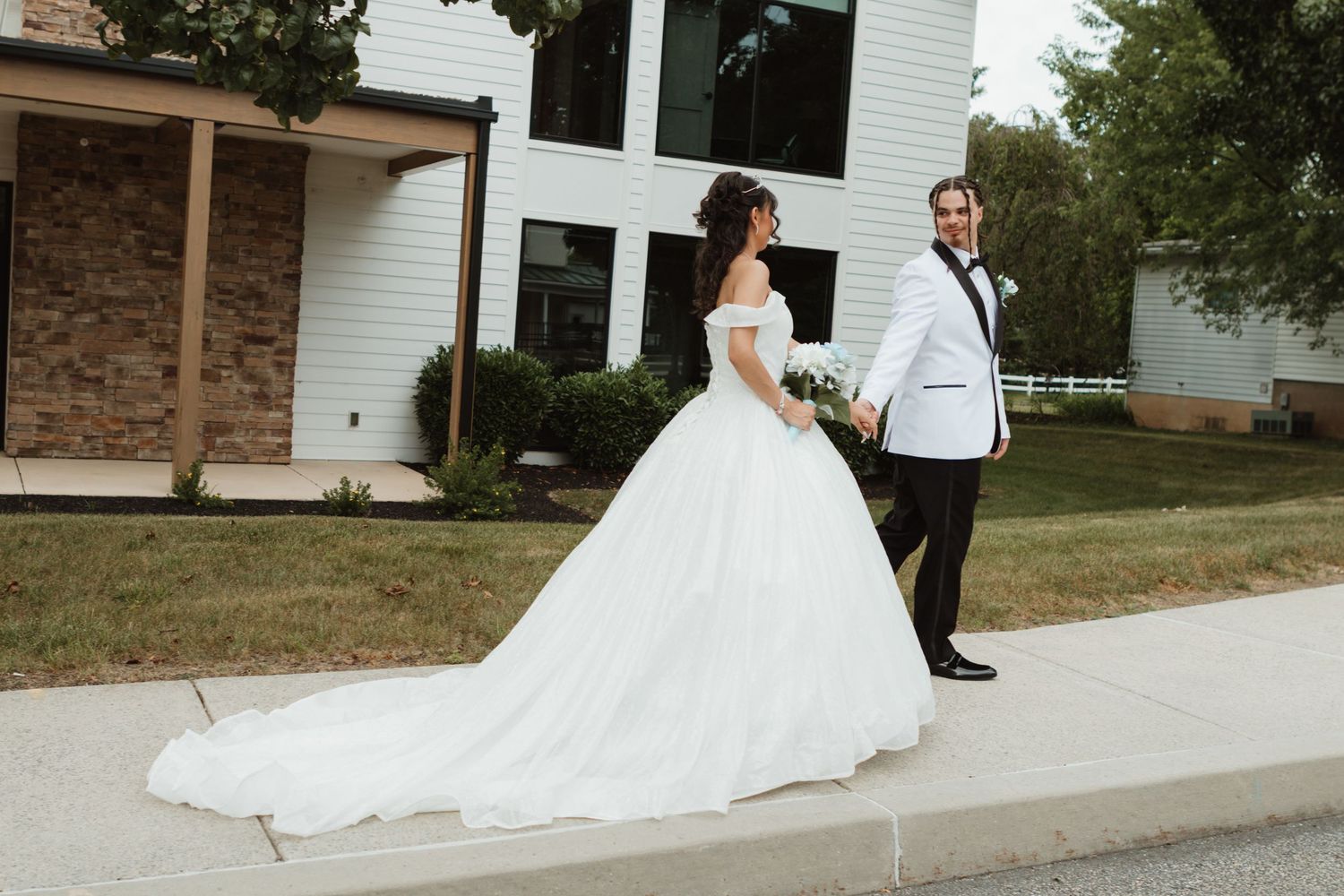 A bride in a white ball gown and groom in black tuxedo walk together on a sidewalk outside a modern white building.