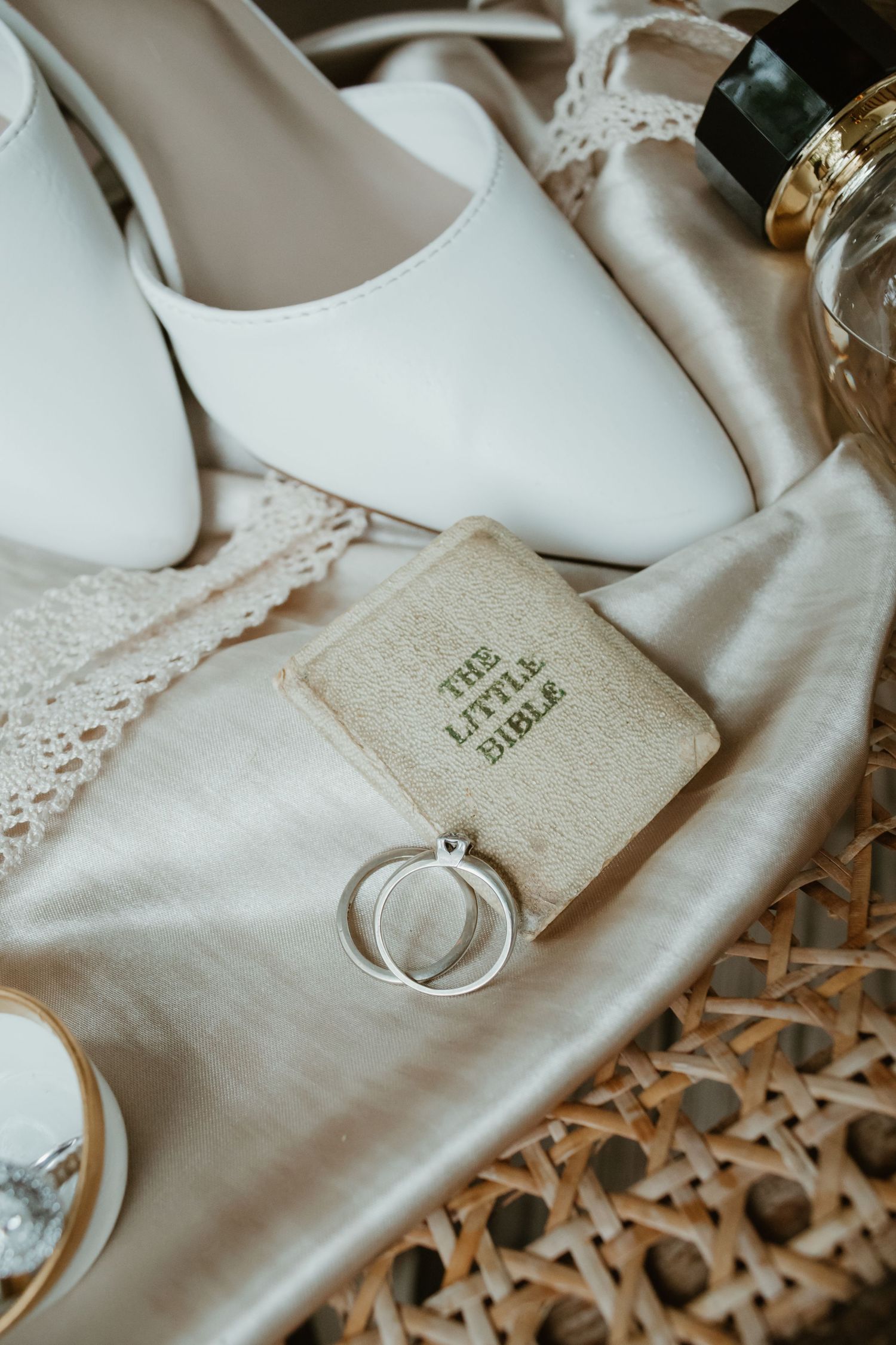 Wedding flat lay with white bridal shoes, rings on a Little Bible, lace fabric, and perfume bottle on wicker surface.