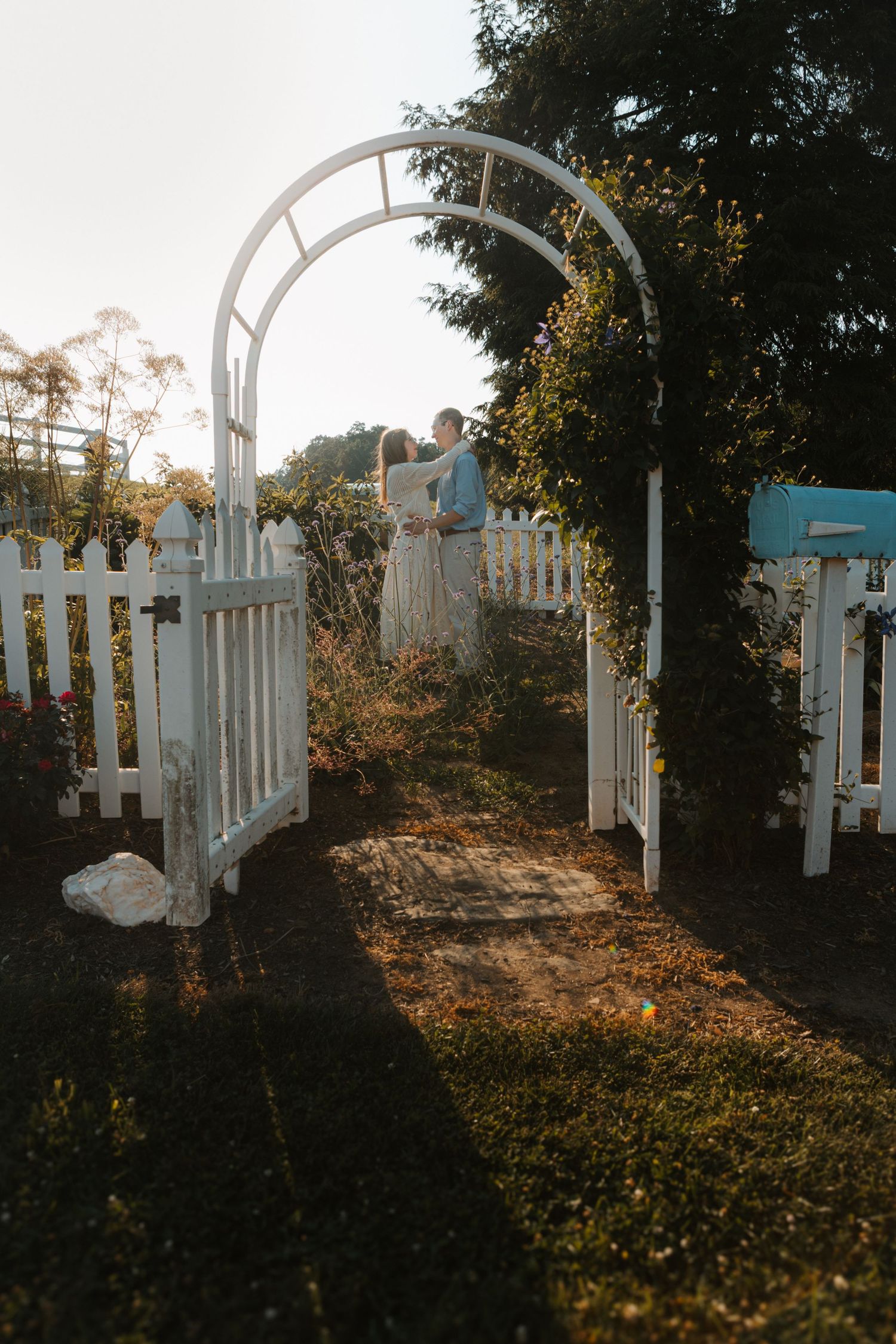 Romantic couple embracing under a white garden arch with picket fence at golden hour sunset.