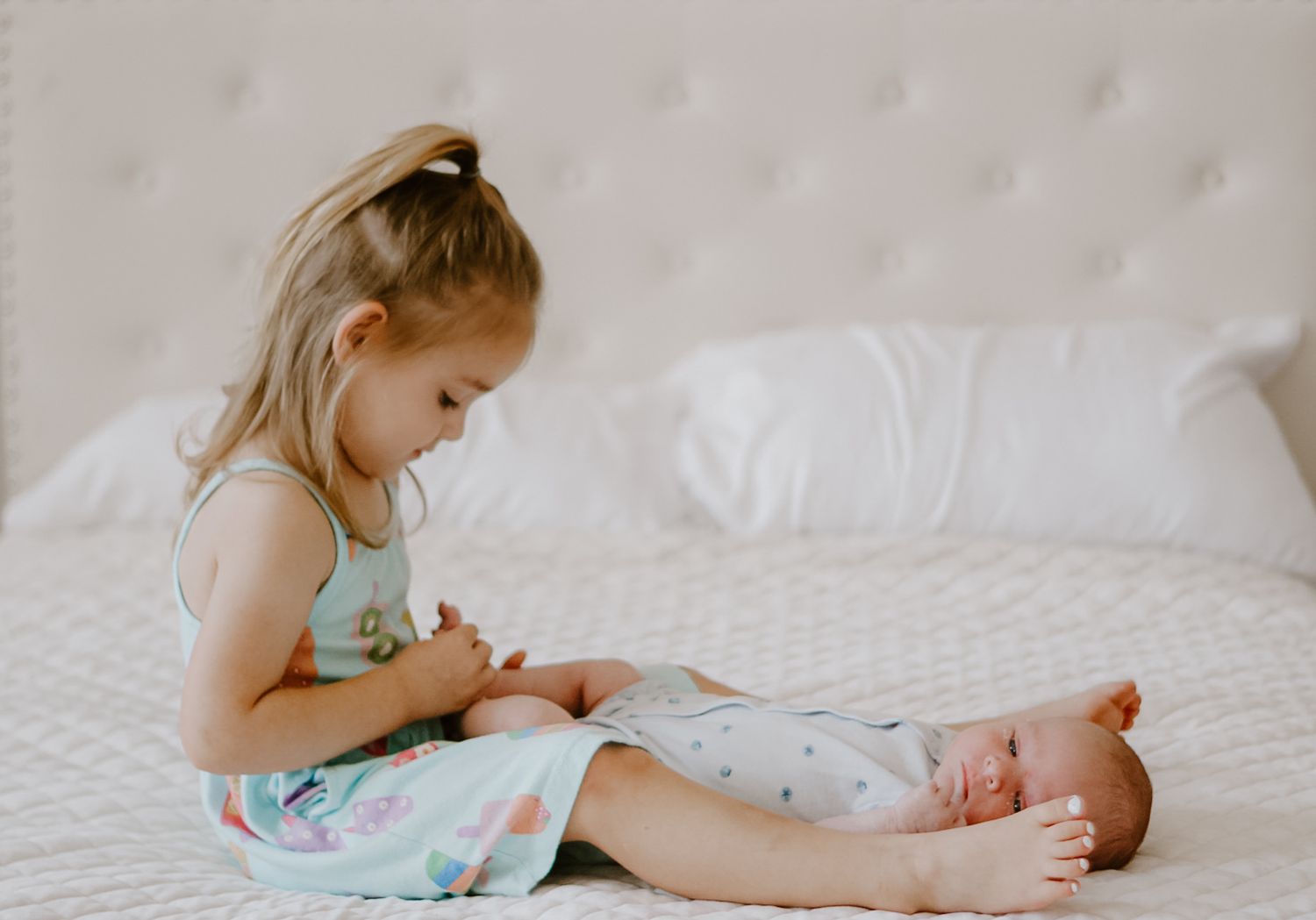 A small child in a turquoise outfit sits on a white bed looking down at a sleeping baby.