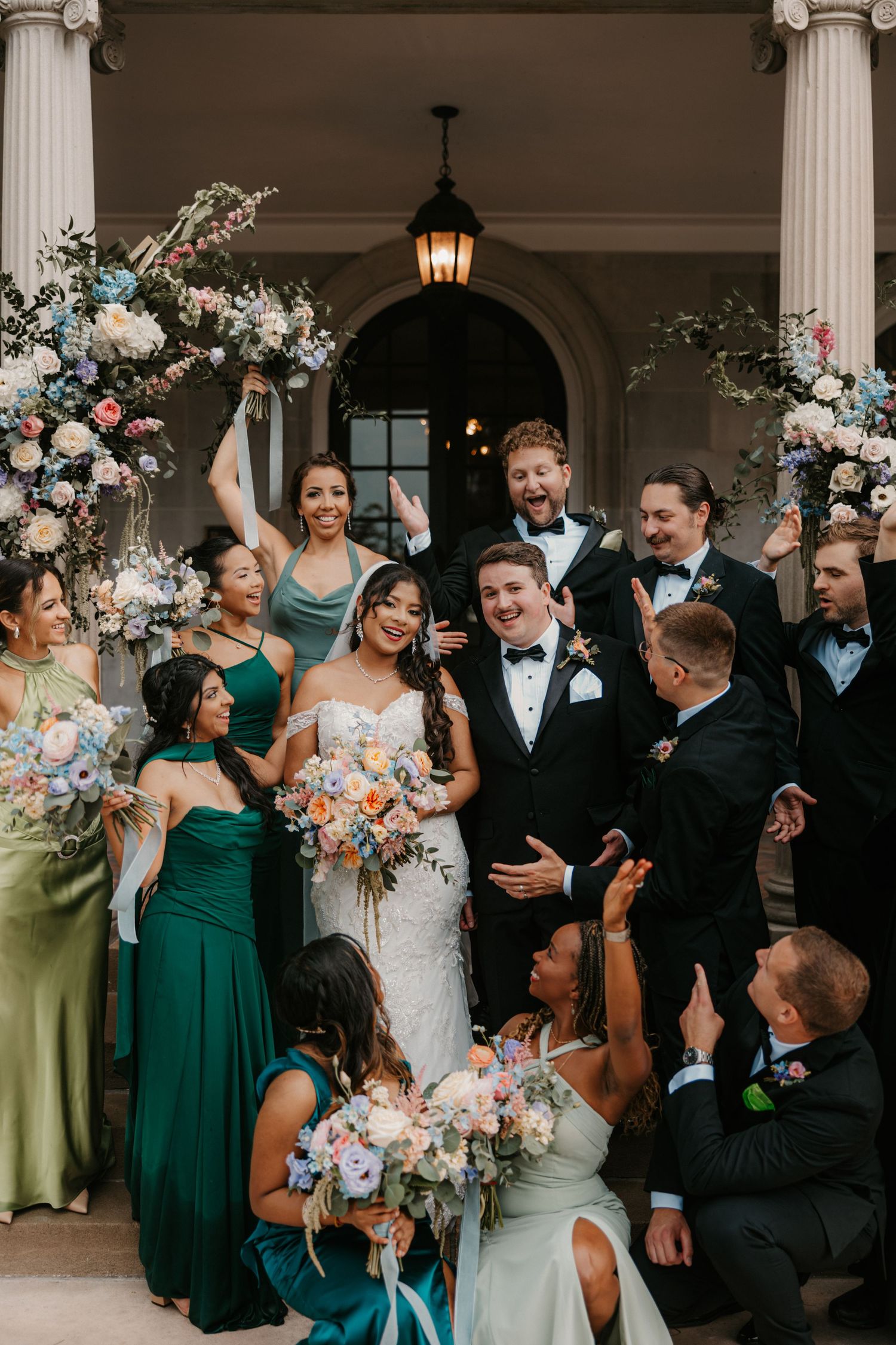 Wedding party celebrates with bride and groom on venue steps, bridesmaids in green dresses holding colorful floral bouquets.