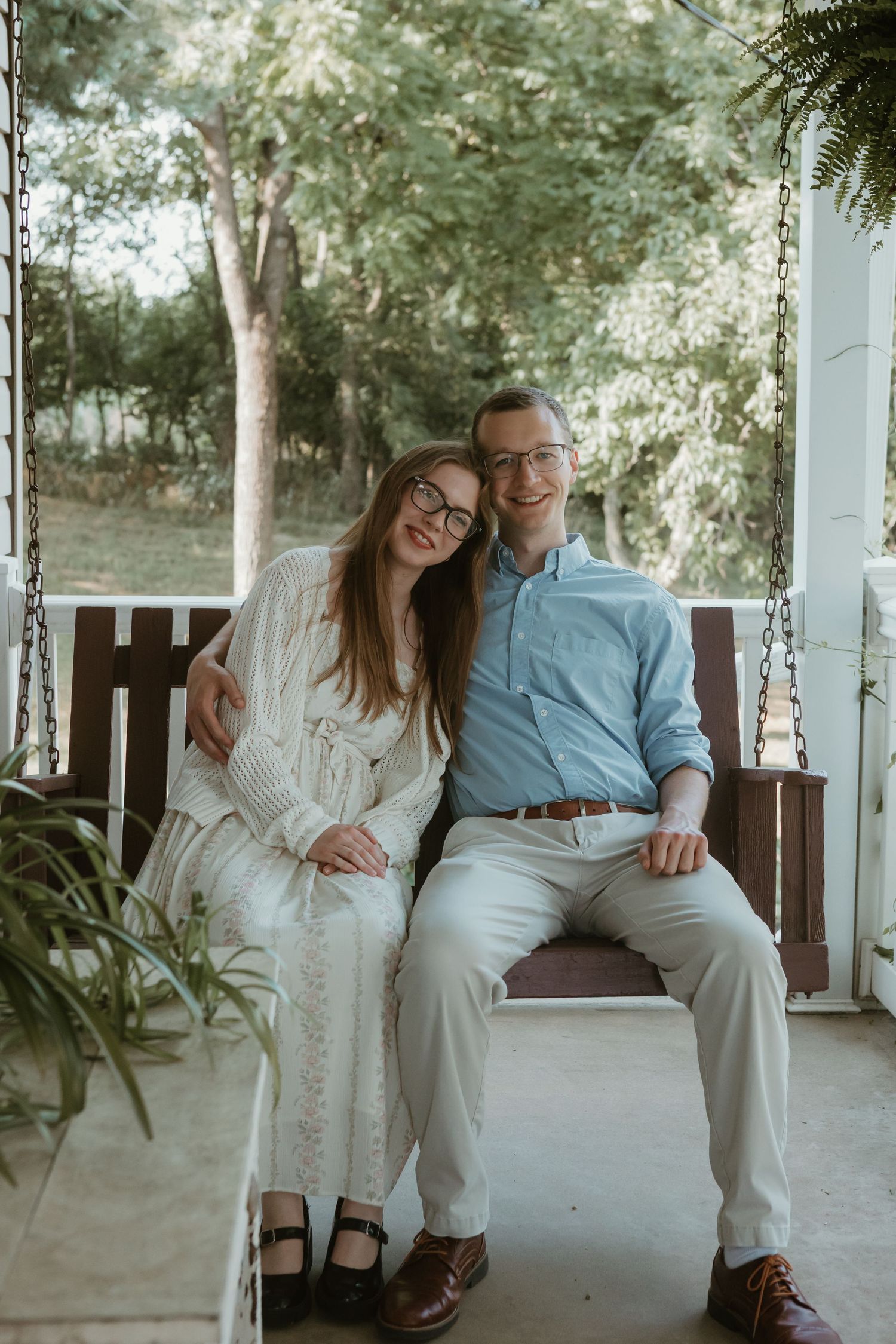 Smiling couple sitting close together on a porch swing, both wearing glasses in a lush garden setting.