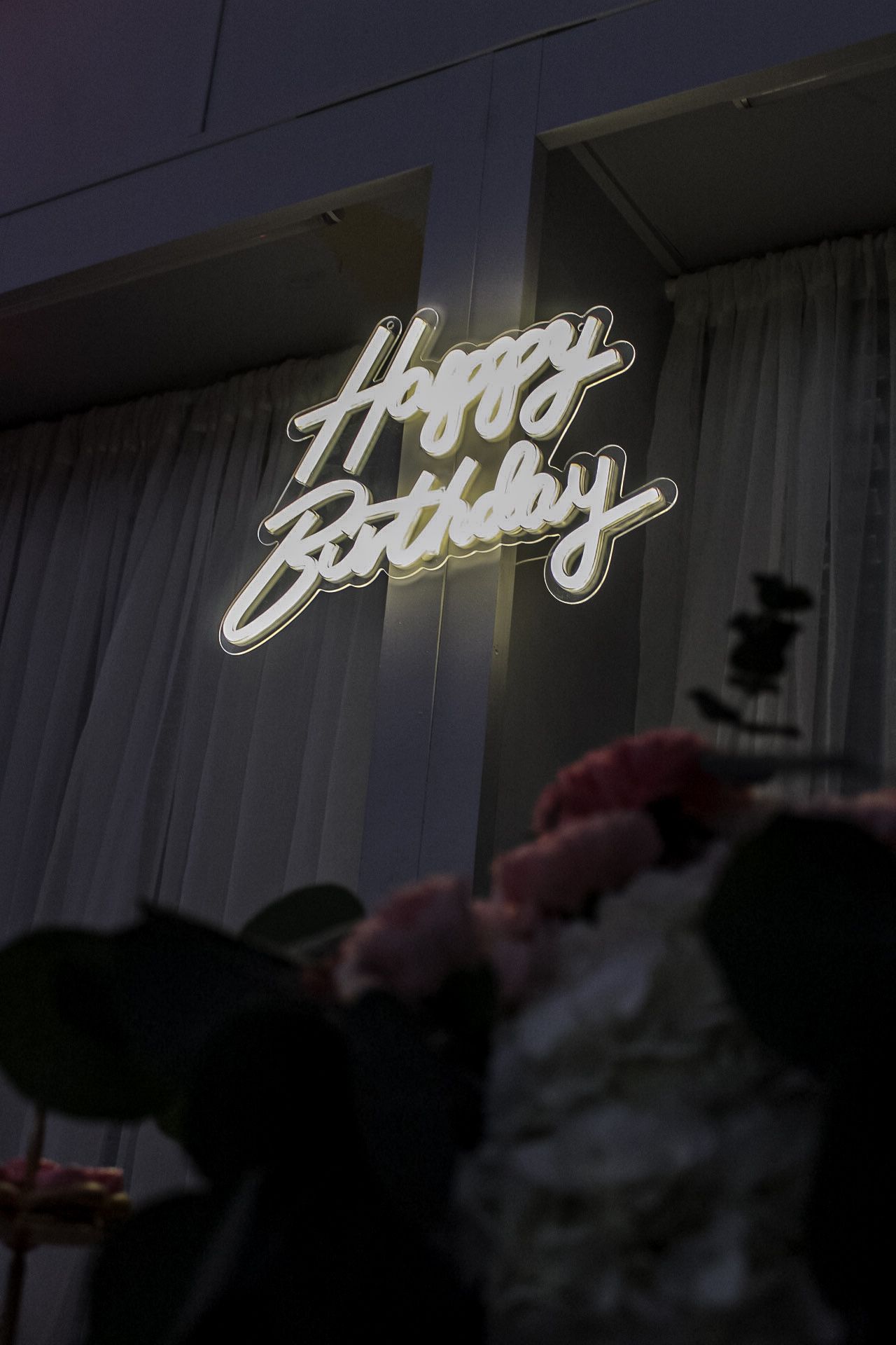 White neon 'Happy Birthday' sign glowing against dark backdrop with floral decorations below.