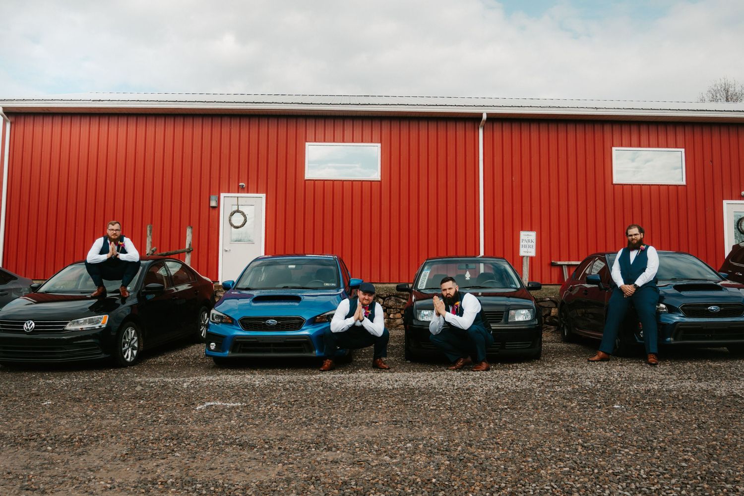 Four groomsmen in vests pose with colorful cars in front of a red barn building on a gravel lot.