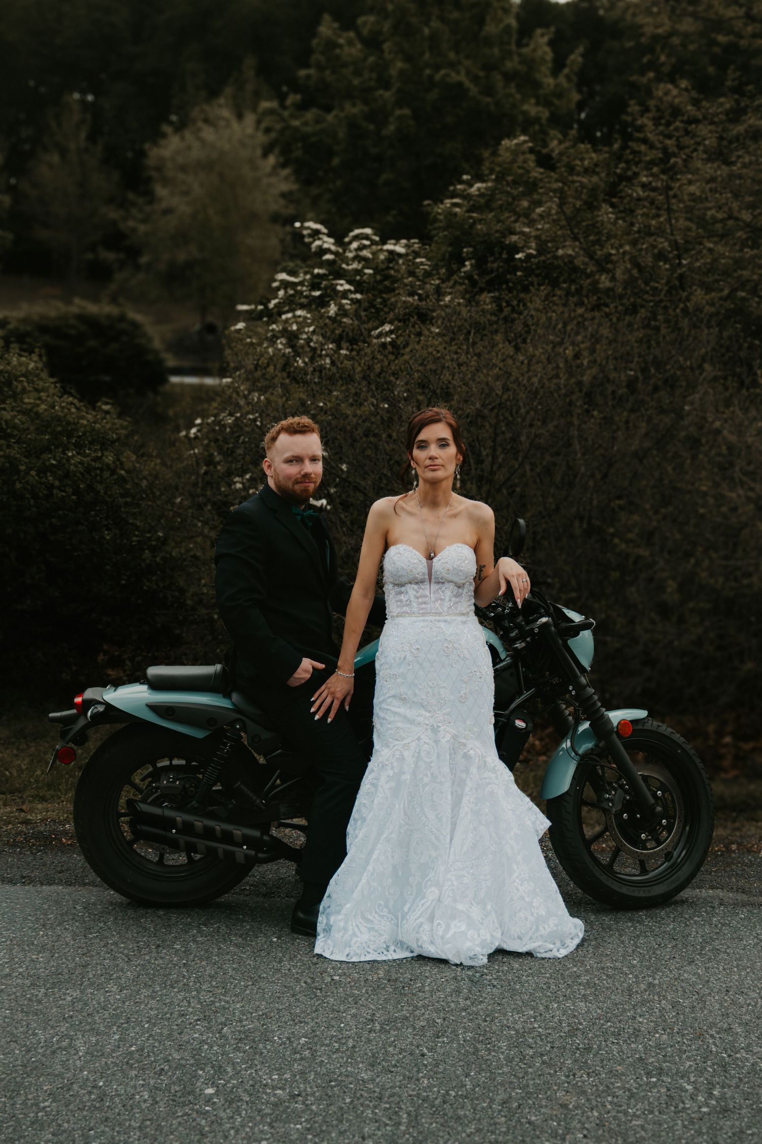 Bride in white lace gown and groom in black suit posing with teal motorcycle outdoors.