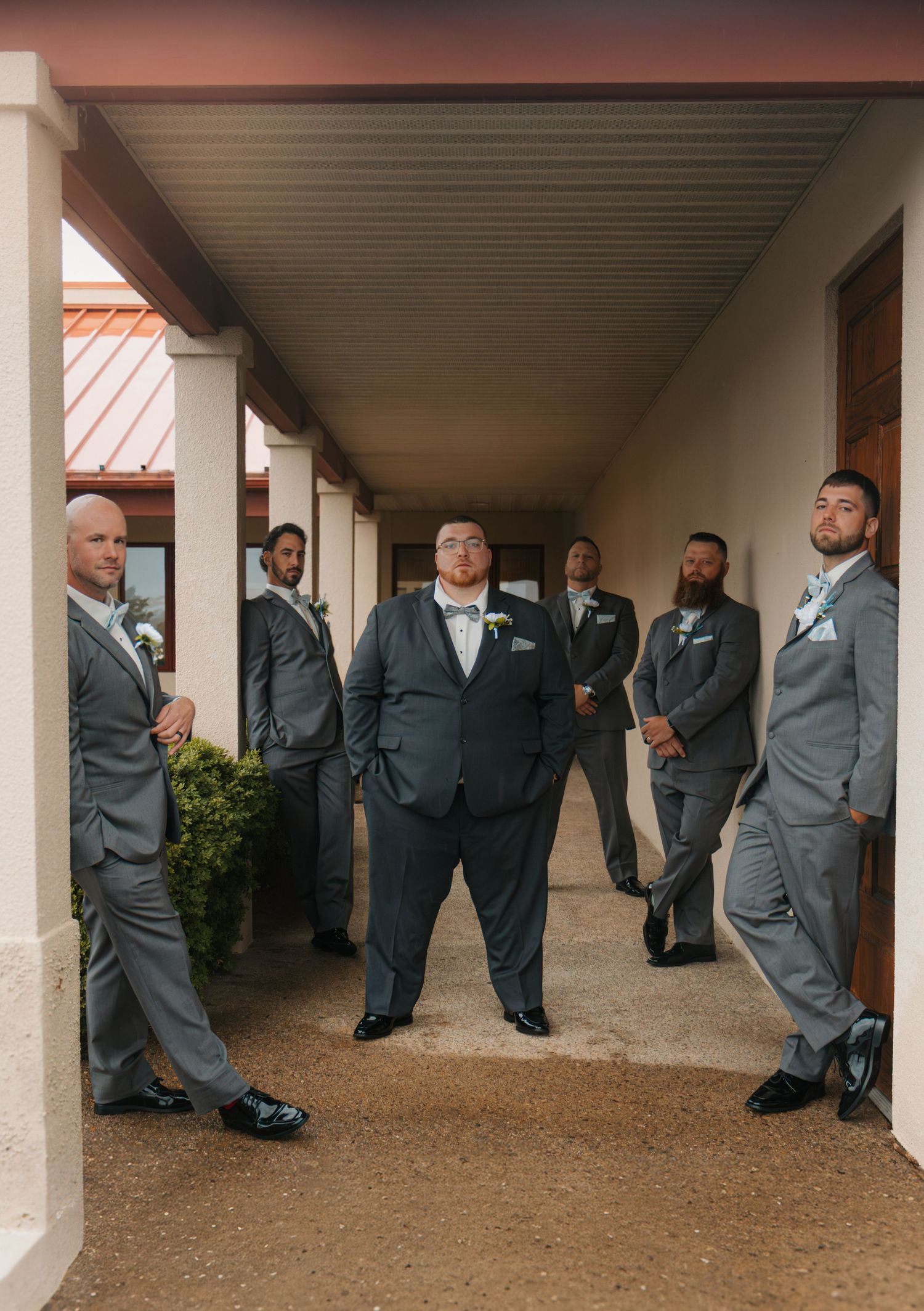 Groom and groomsmen posing in grey suits with bow ties in a covered outdoor corridor at a wedding venue.