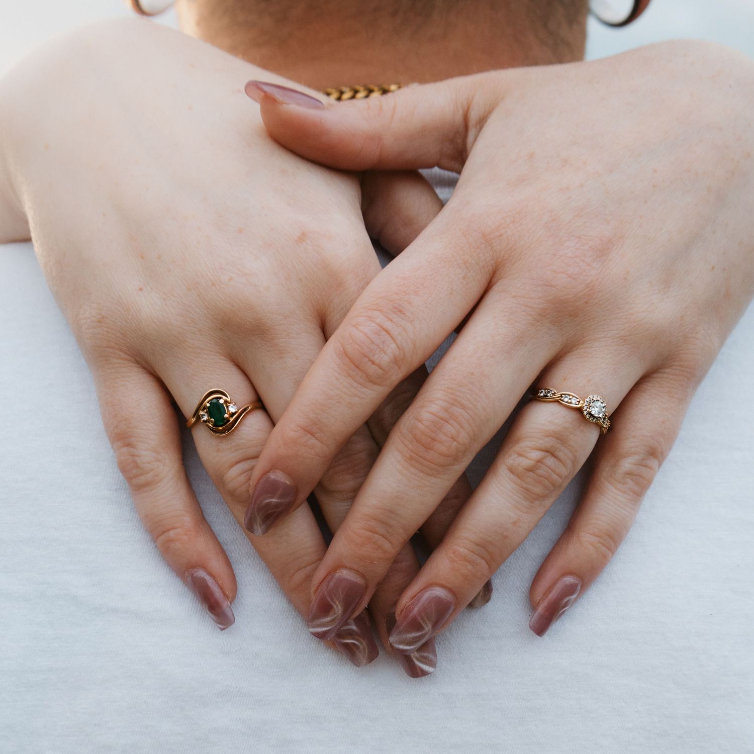 Close-up of two hands adorned with gold rings featuring green stone and chain designs, with neutral manicured nails.
