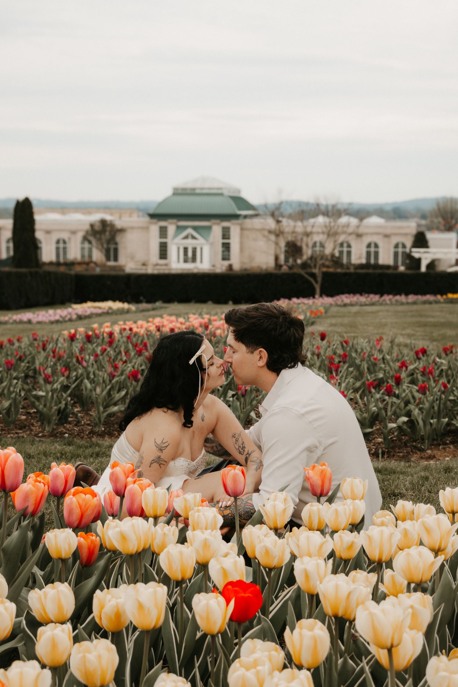 Couple sitting among colorful tulips in a garden, sharing a romantic moment with a historic building in the background.
