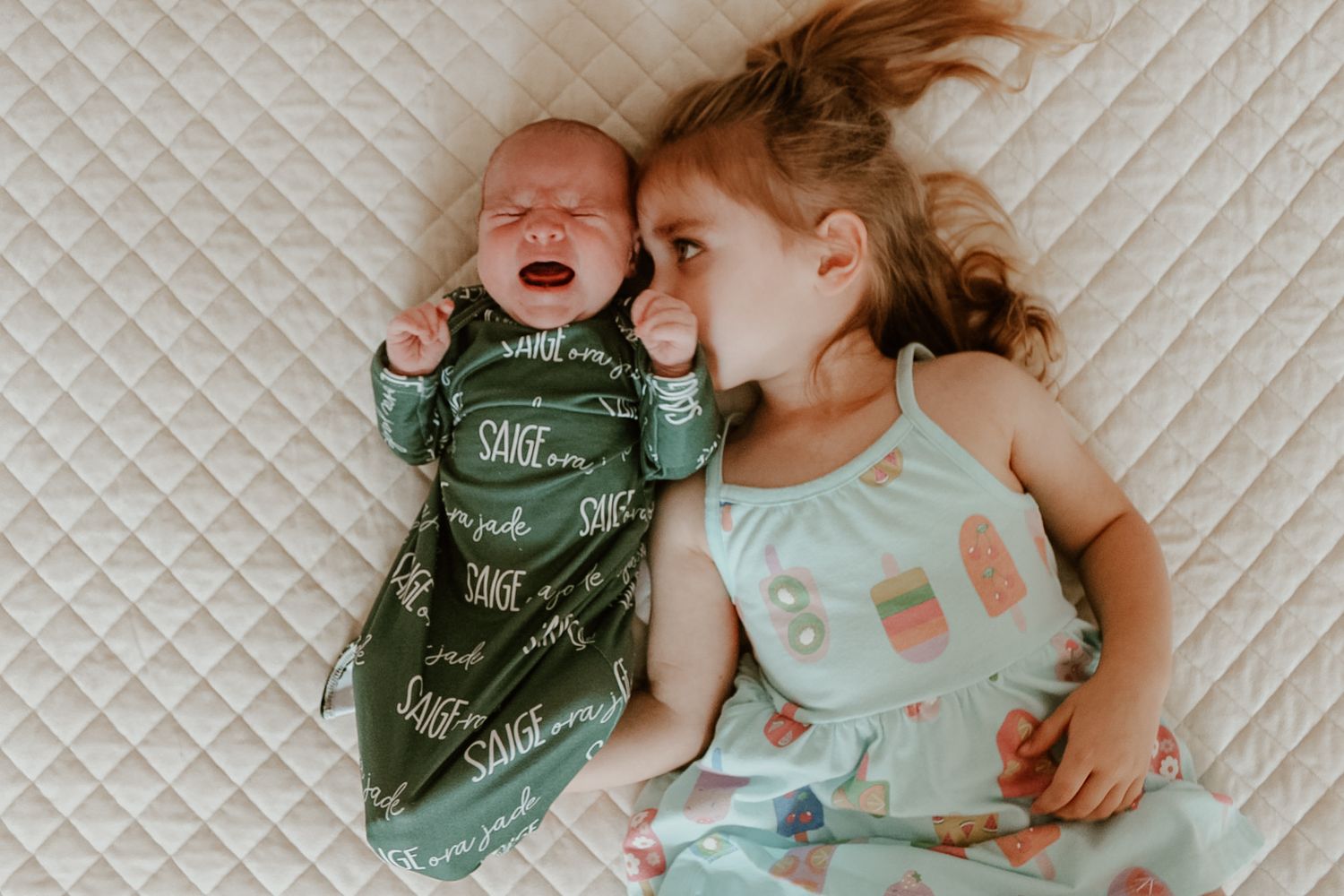 Two babies lying on quilted white bedding wearing mint green and bandana outfits.