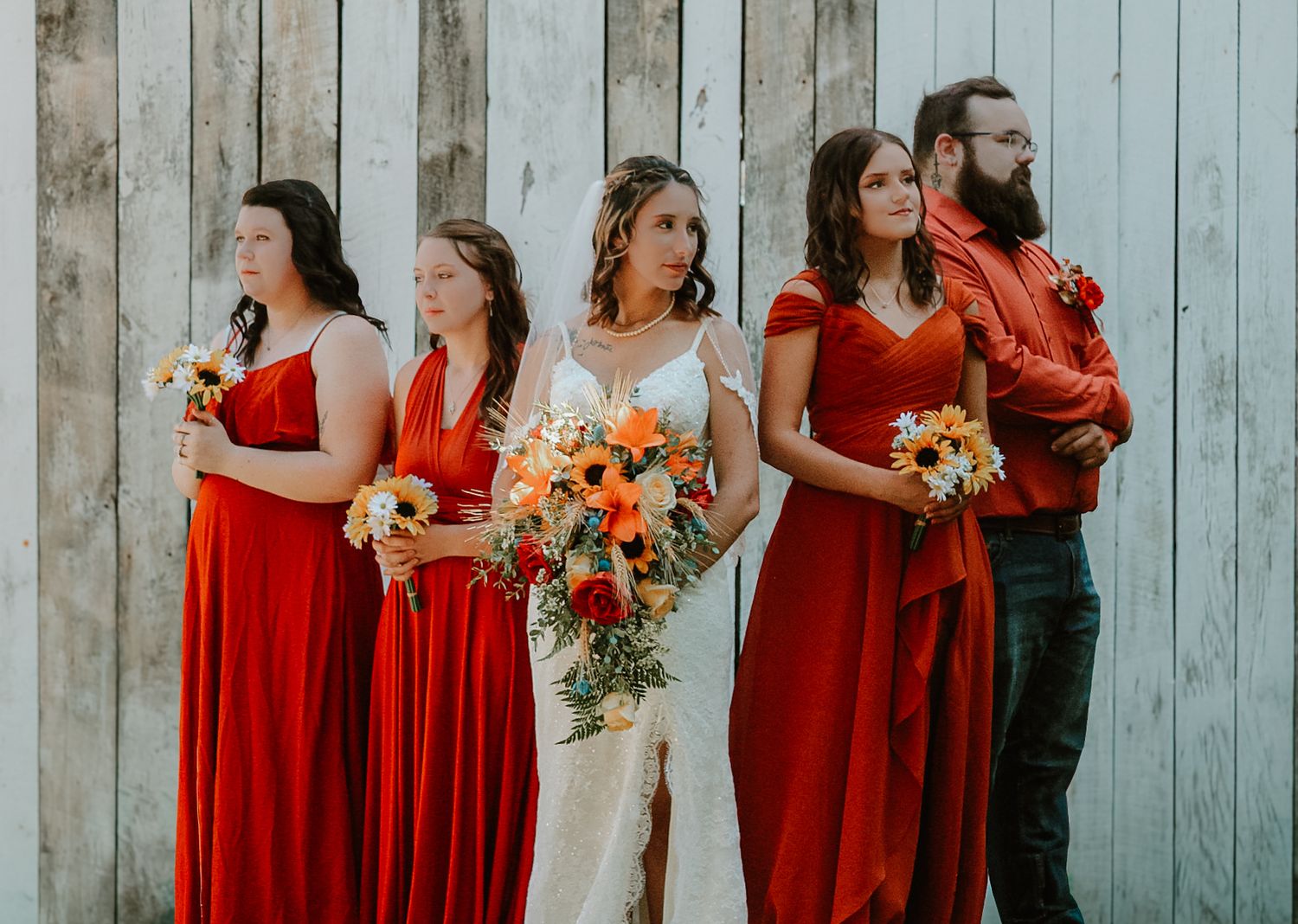 Wedding party in vibrant red dresses and a white gown pose against rustic blue wooden wall with orange bouquets.