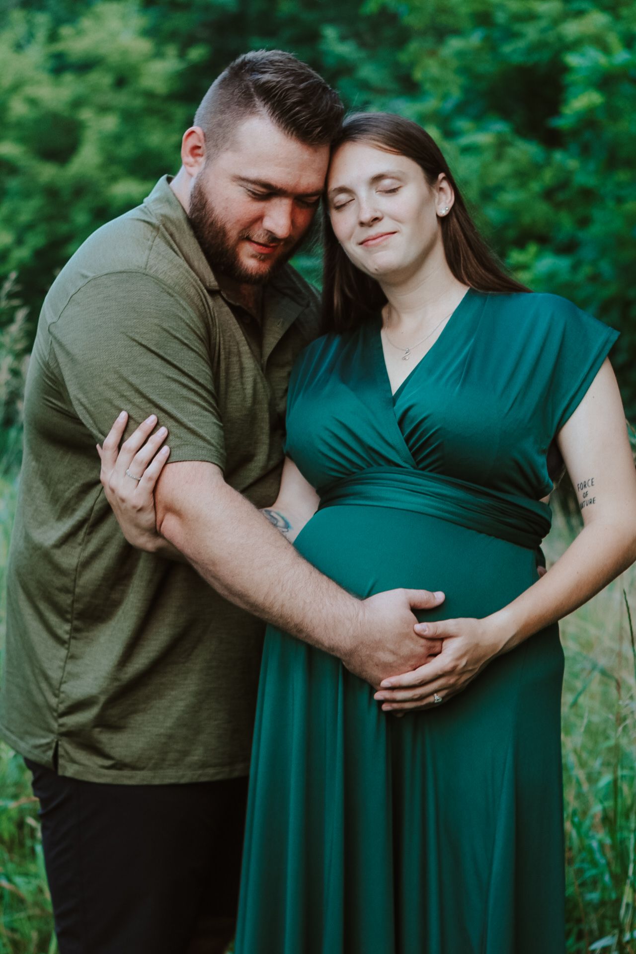Tender maternity portrait of couple in green dress and olive shirt against forest backdrop.
