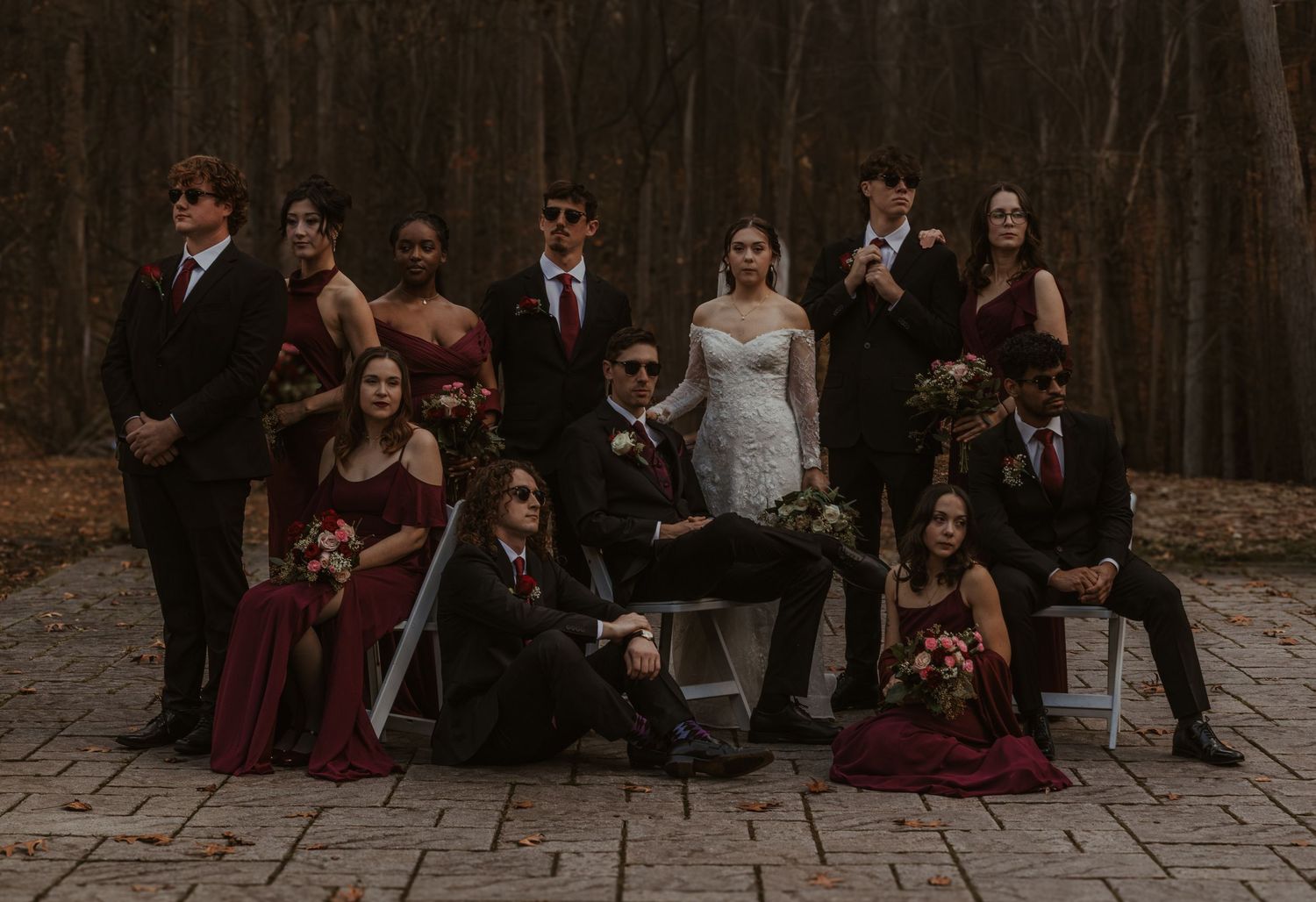 A wedding party poses together in formal burgundy and black attire against a moody forest backdrop.