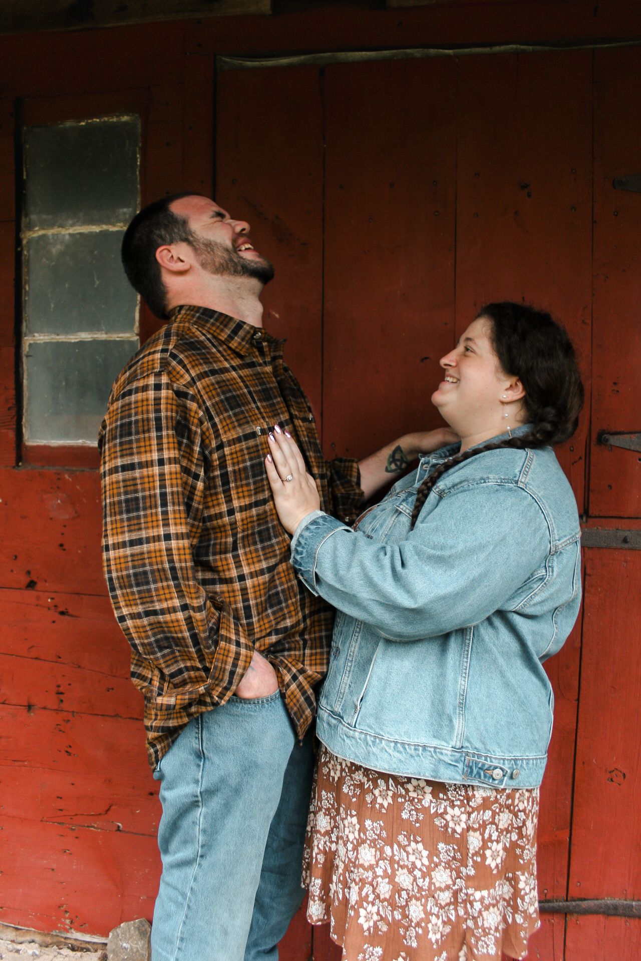Joyful couple laughing against red barn wall in casual autumn outfits.