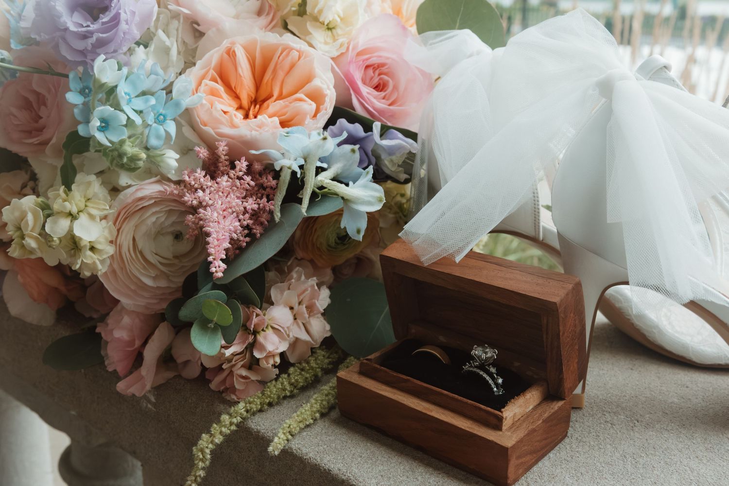 Pastel wedding bouquet with roses beside a wooden ring box with diamond rings and white bridal heels.
