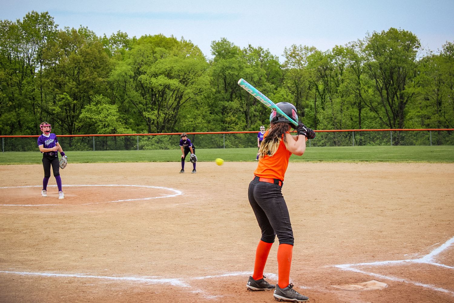 Softball batter in orange jersey takes stance at home plate during game.