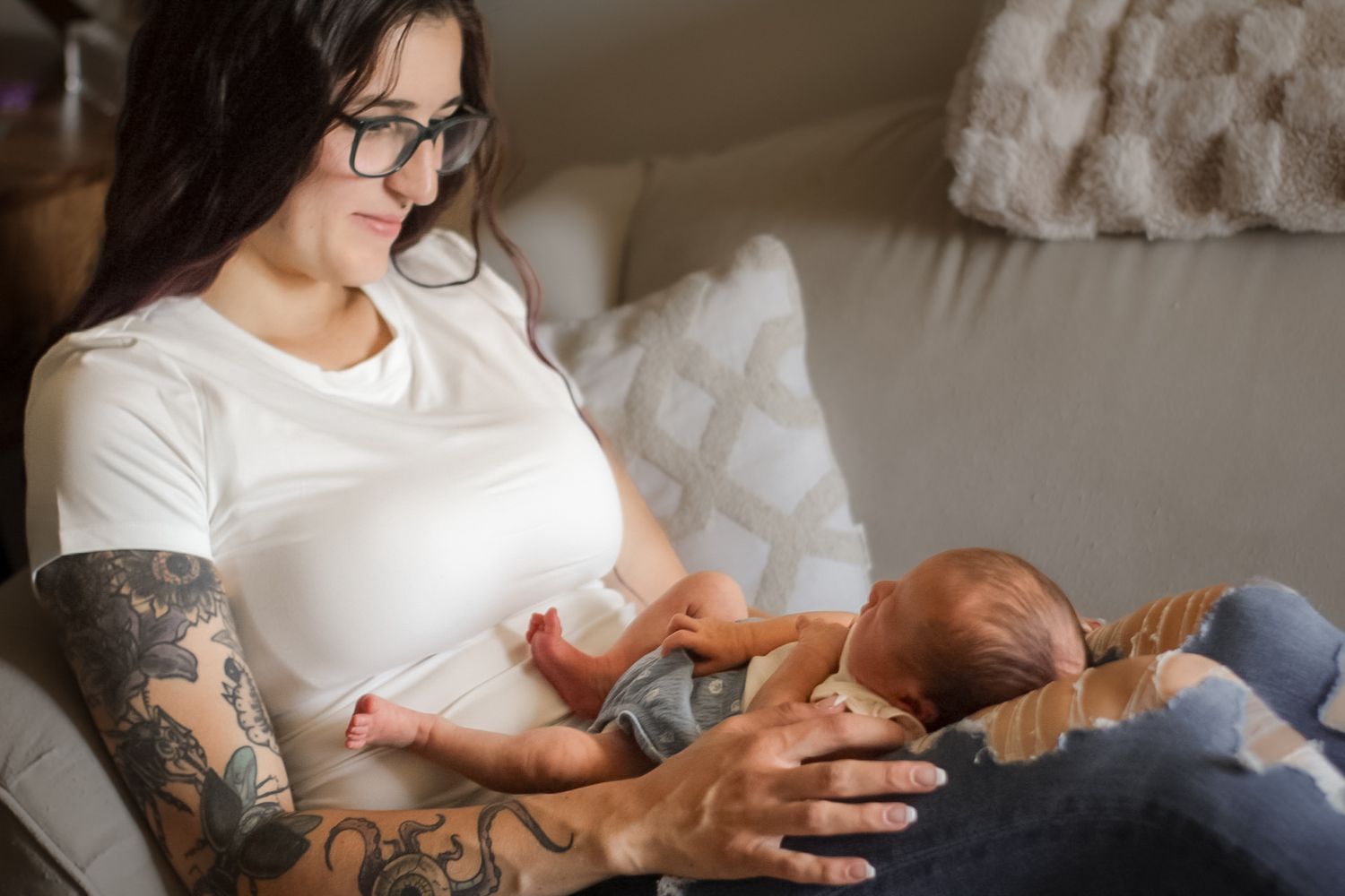 Tattooed parent in white shirt tenderly caring for infant on couch.