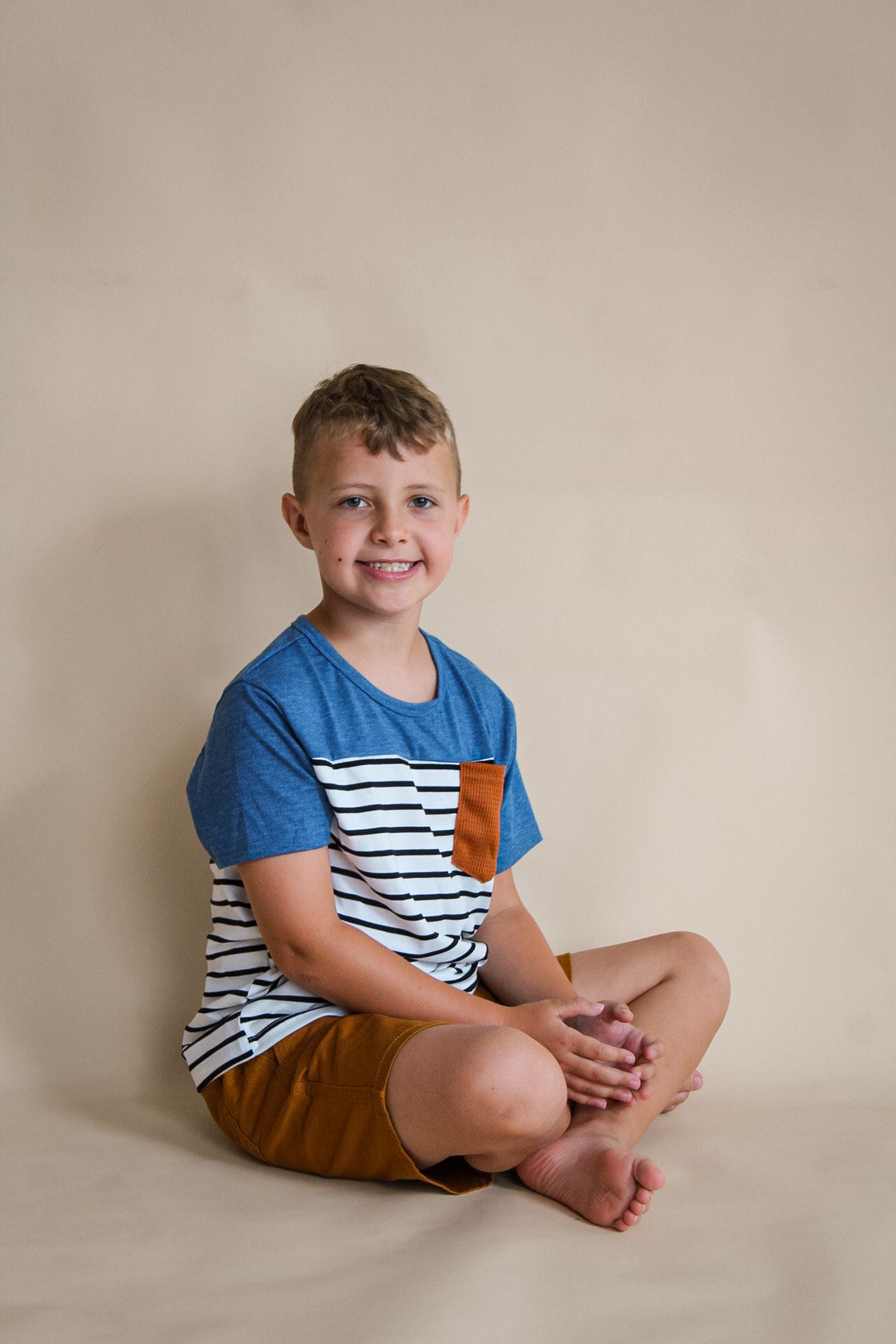 A young child sits cross-legged on a beige backdrop wearing a blue and white striped t-shirt with brown shorts.
