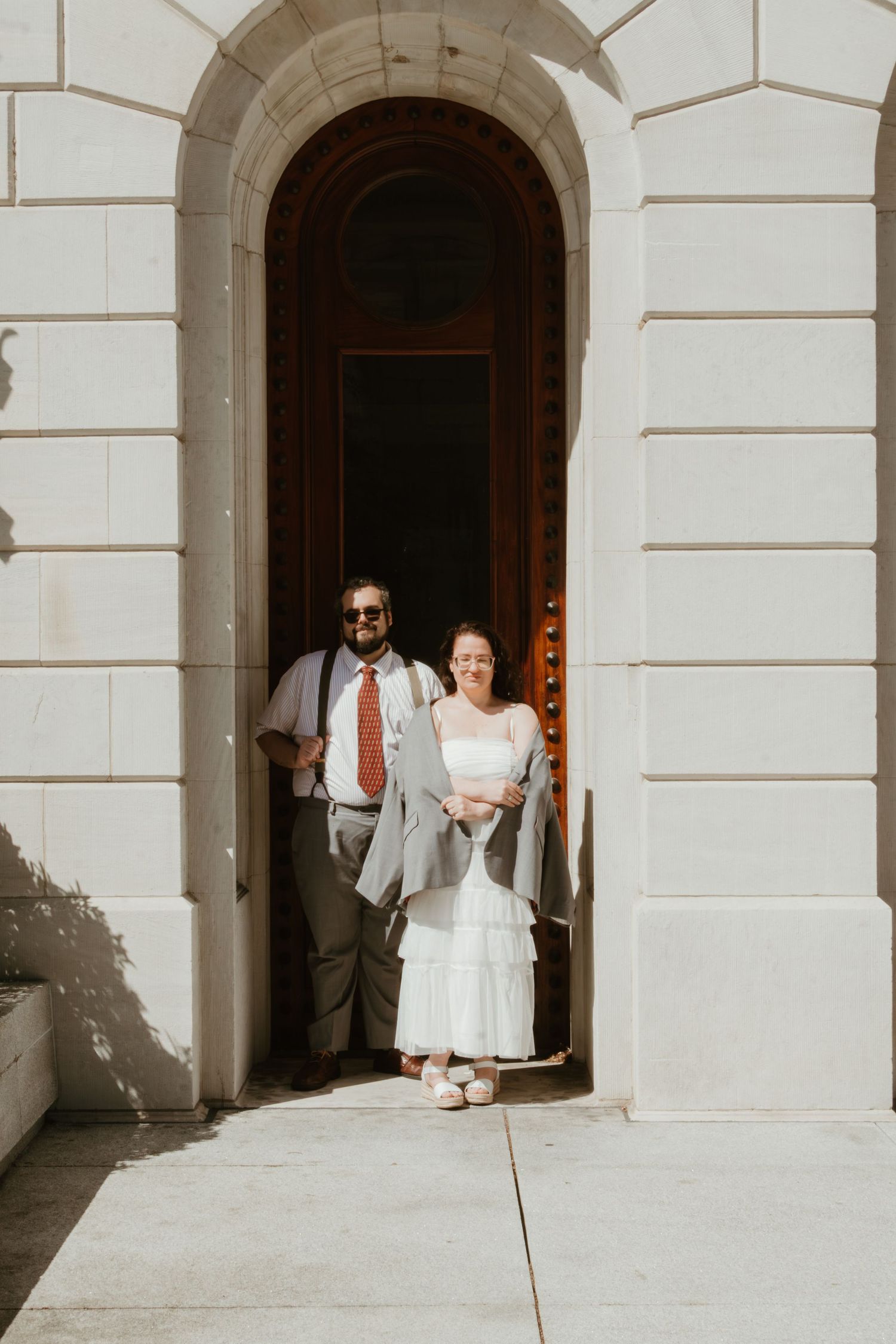 Couple in wedding attire standing in an arched doorway of a white stone building on their wedding day.