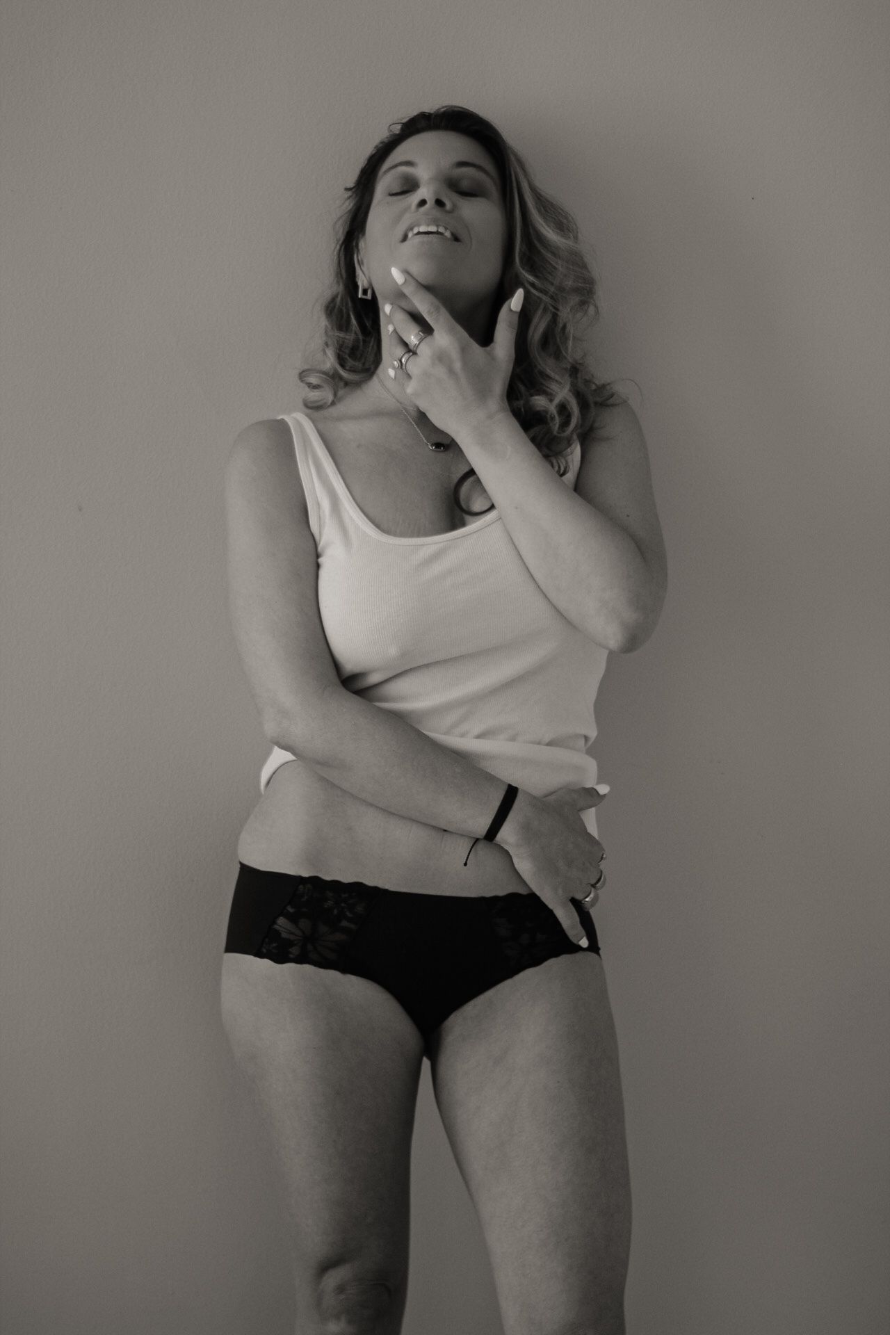 Black and white boudoir portrait of woman in tank top and underwear against neutral wall.