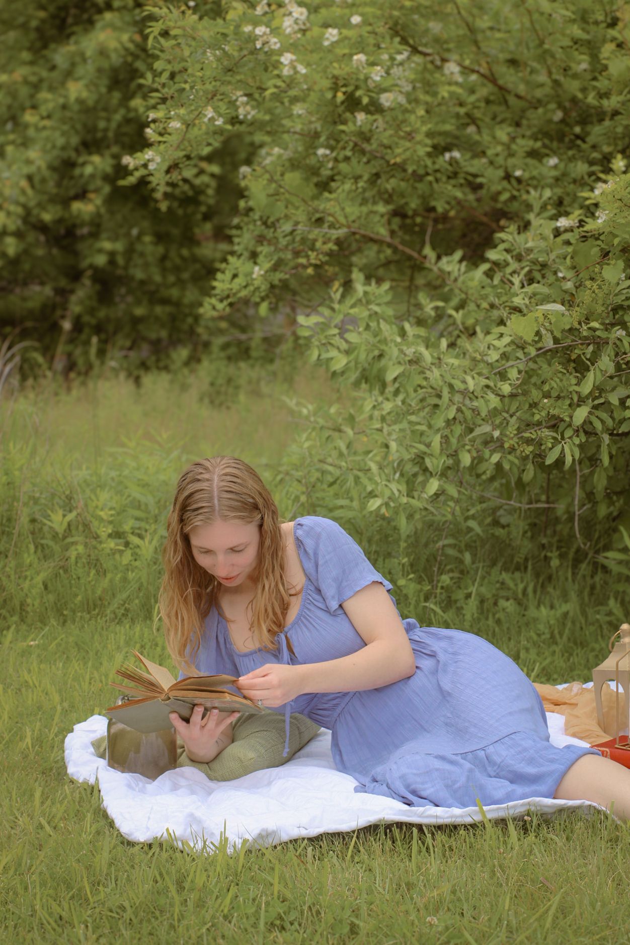 Woman in a blue dress enjoys a picnic on a white blanket with lanterns and old vintage books. Cottage core vibes