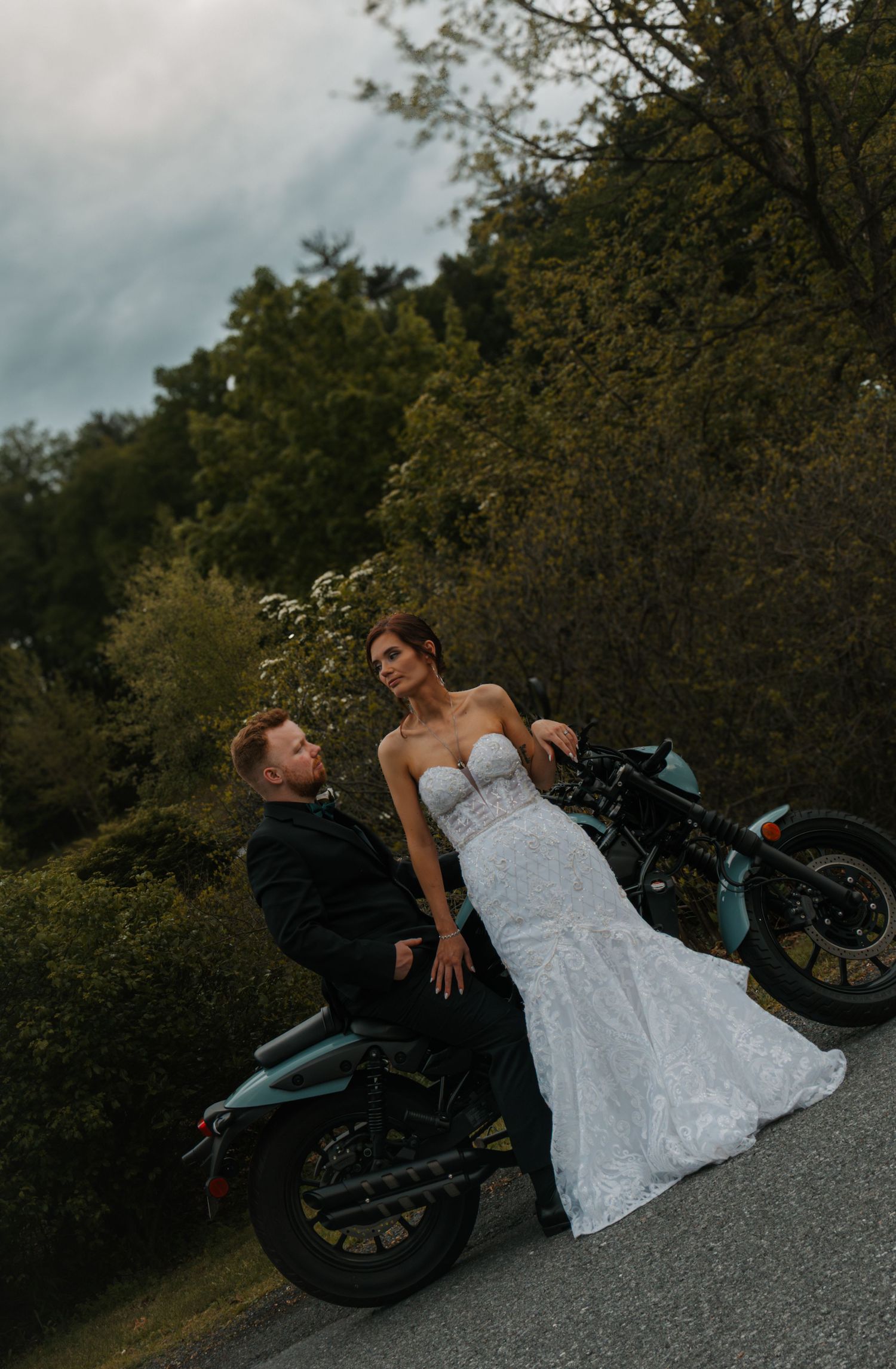 Bride in strapless lace gown and groom in black suit posing romantically beside a motorcycle outdoors.