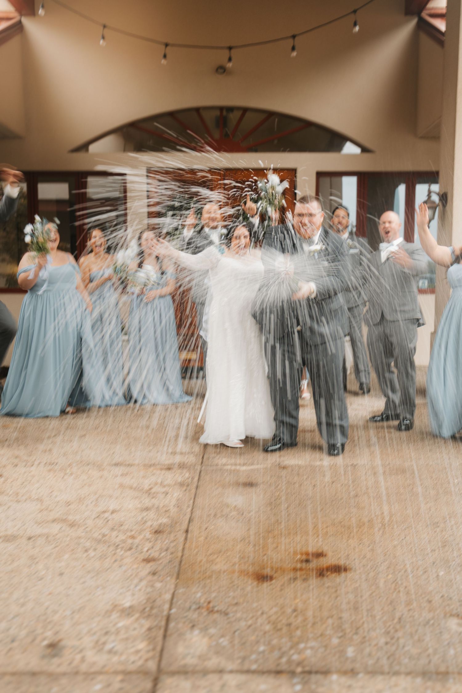 Bride and groom celebrate with champagne spray as wedding party cheers in dusty blue and gray attire.