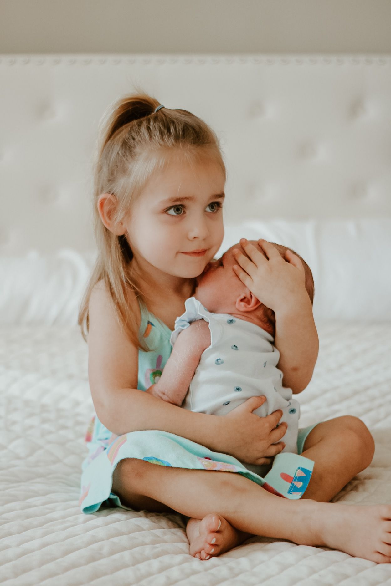 A young child in a mint green dress holds and gazes lovingly at a newborn baby on a white textured bed.