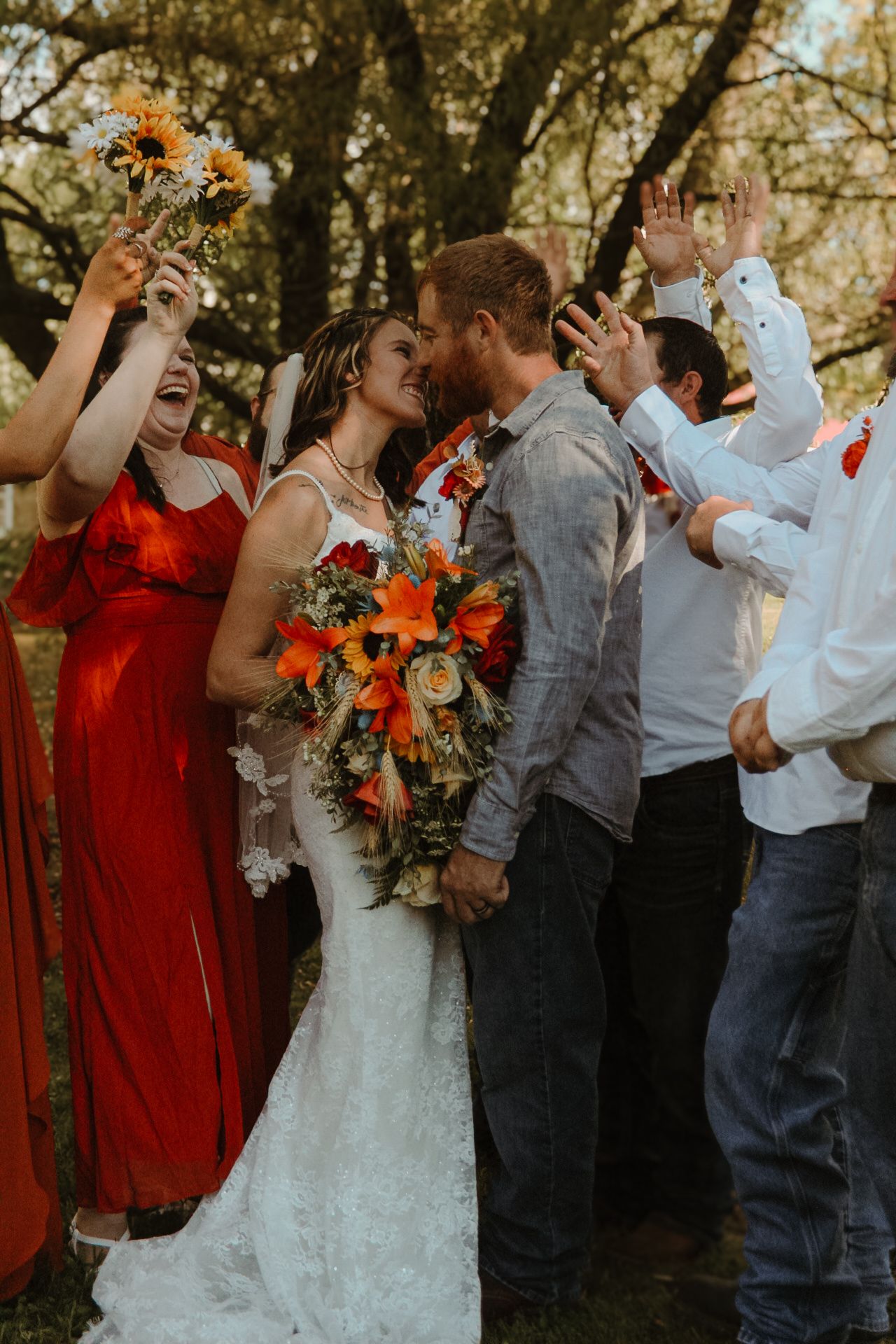 Wedding party in red dresses and casual attire poses together under autumn trees with orange foliage.