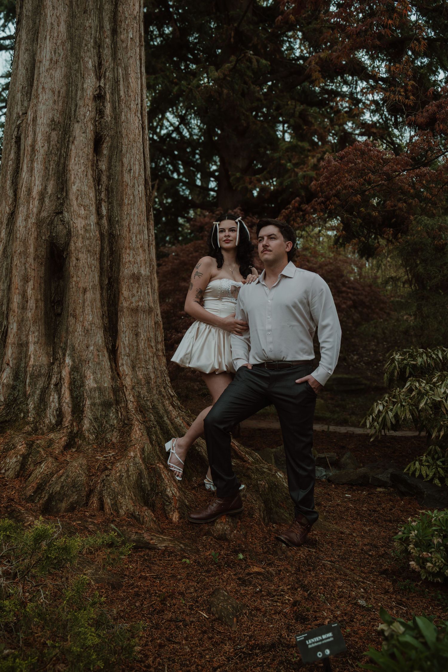 Couple posing by a large tree in a forest garden, woman in white dress with floral headpiece, man in white shirt.