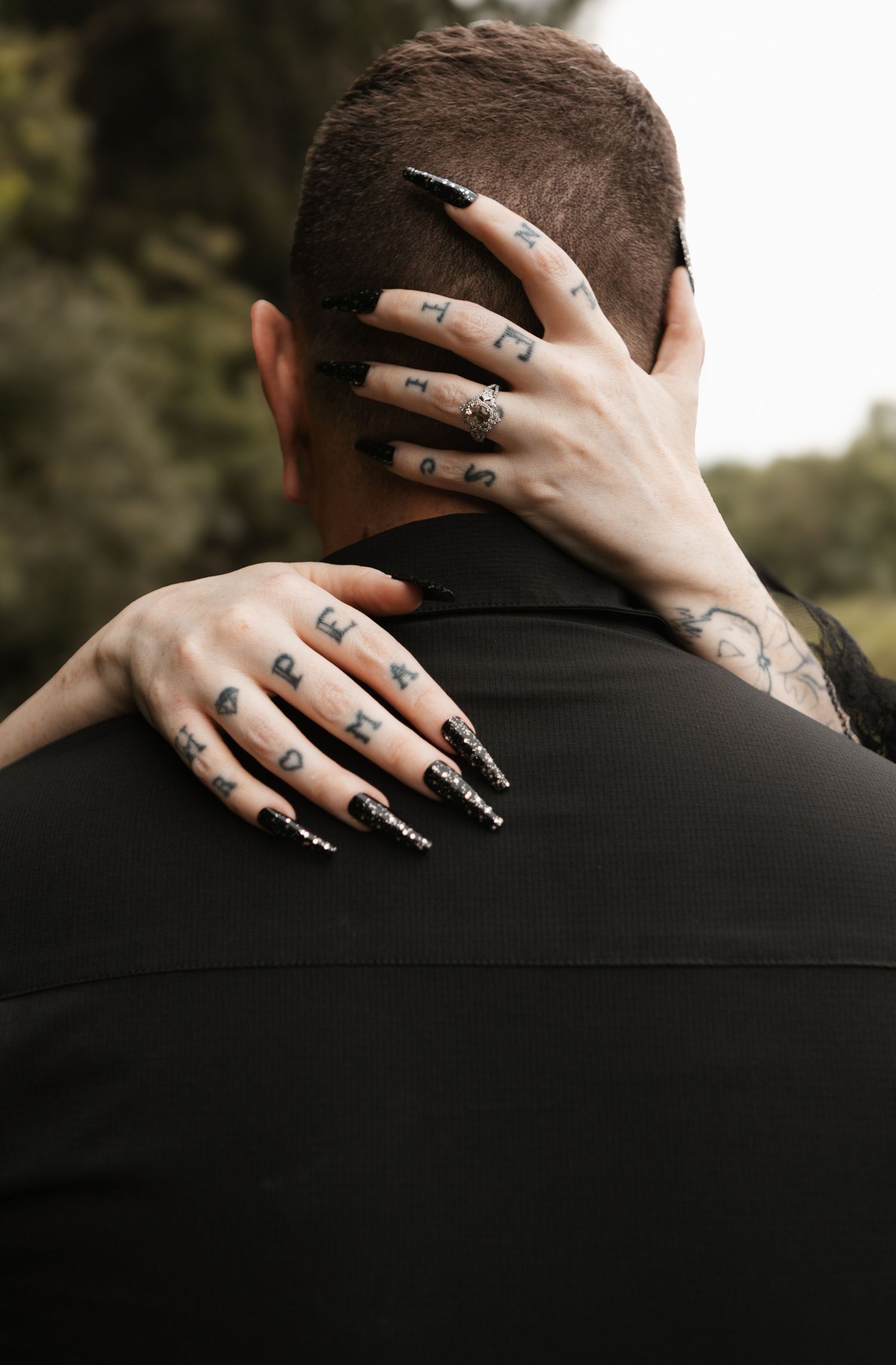 Person with tattooed fingers and black stiletto nails embracing someone from behind, wearing a black shirt outdoors.
