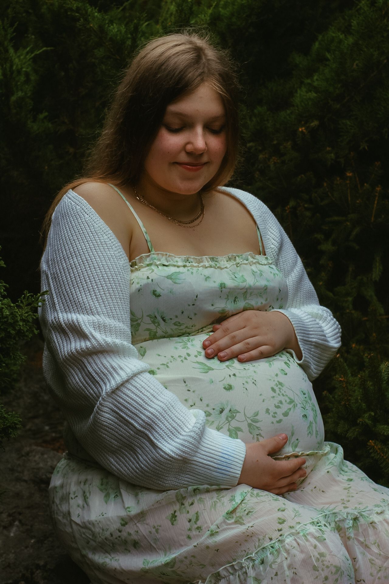 A serene maternity portrait featuring a floral dress and white knit cardigan in a forest setting.