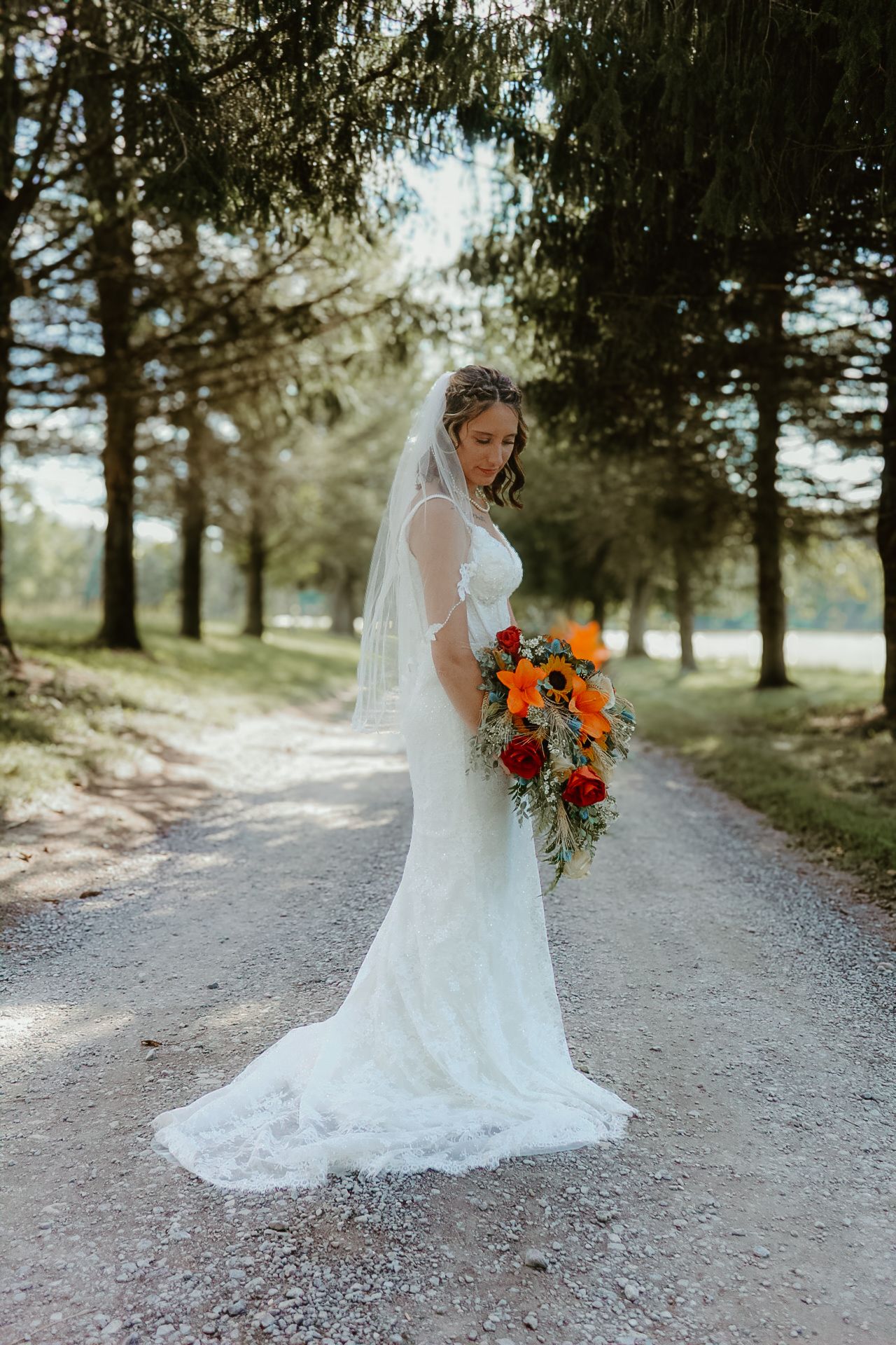 Bride in flowing white gown holds vibrant autumn bouquet while walking down tree-lined path.