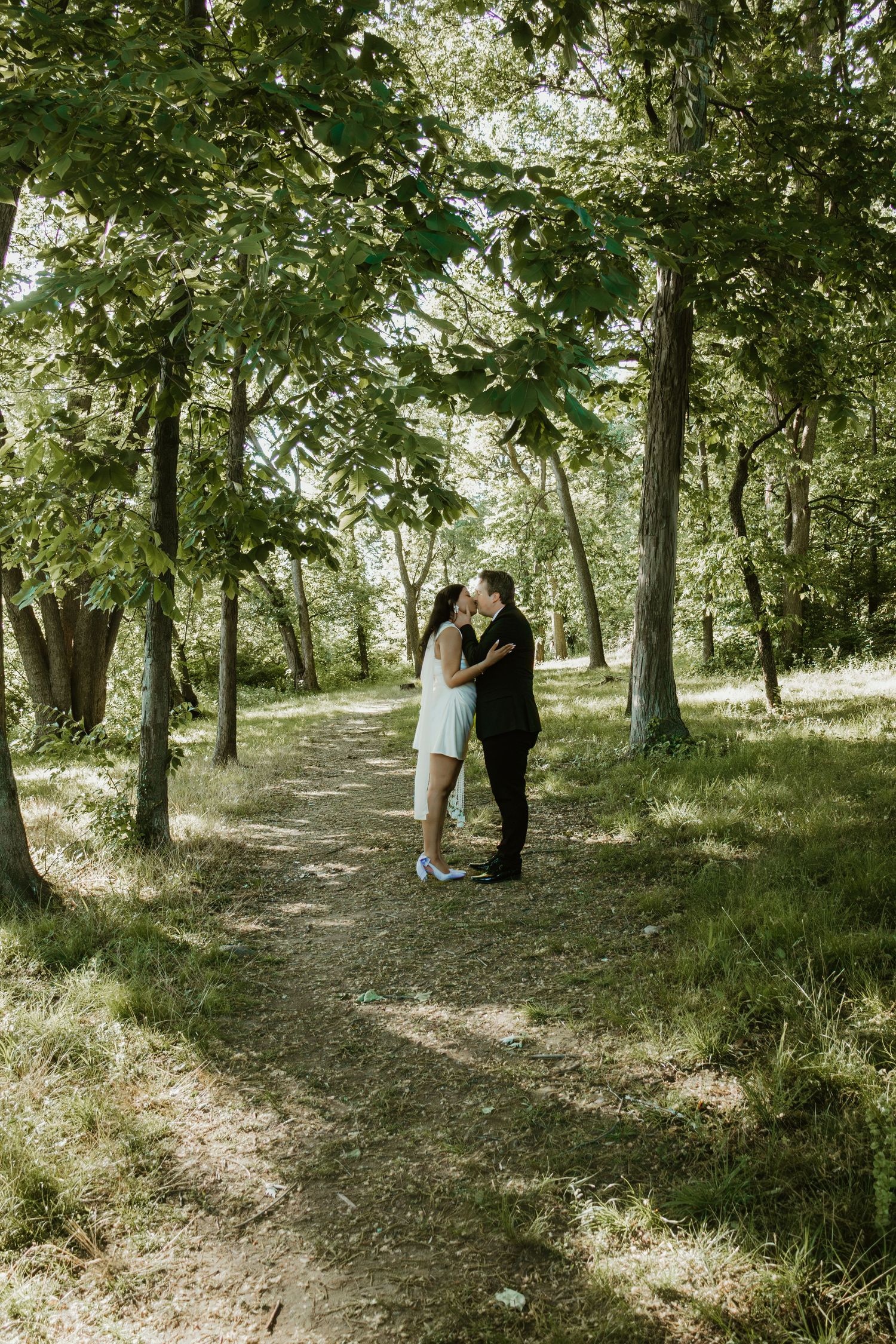 Couple kissing on a sunlit forest path surrounded by lush green trees.