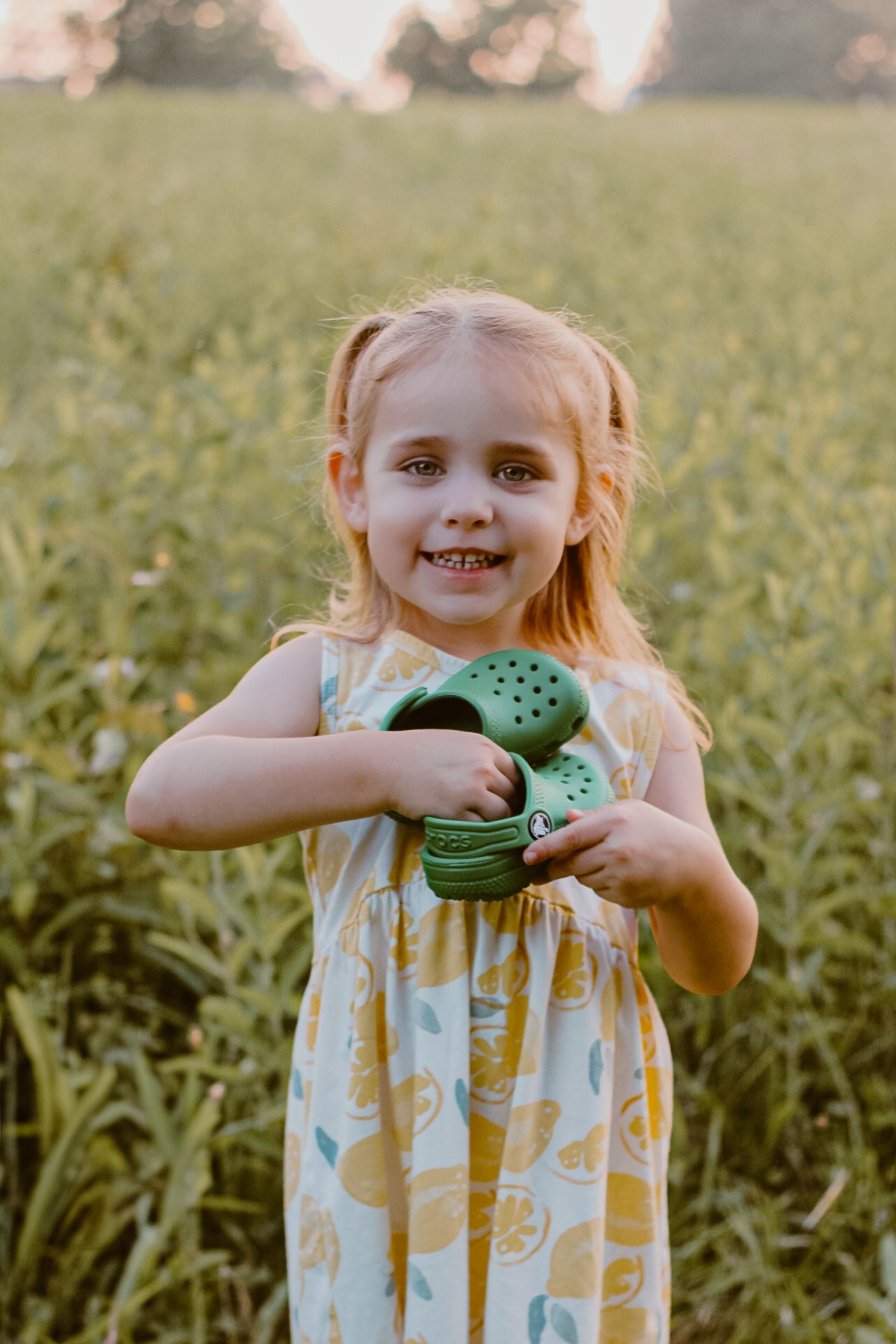 Child in yellow summer dress holding green watering can in field during golden hour.