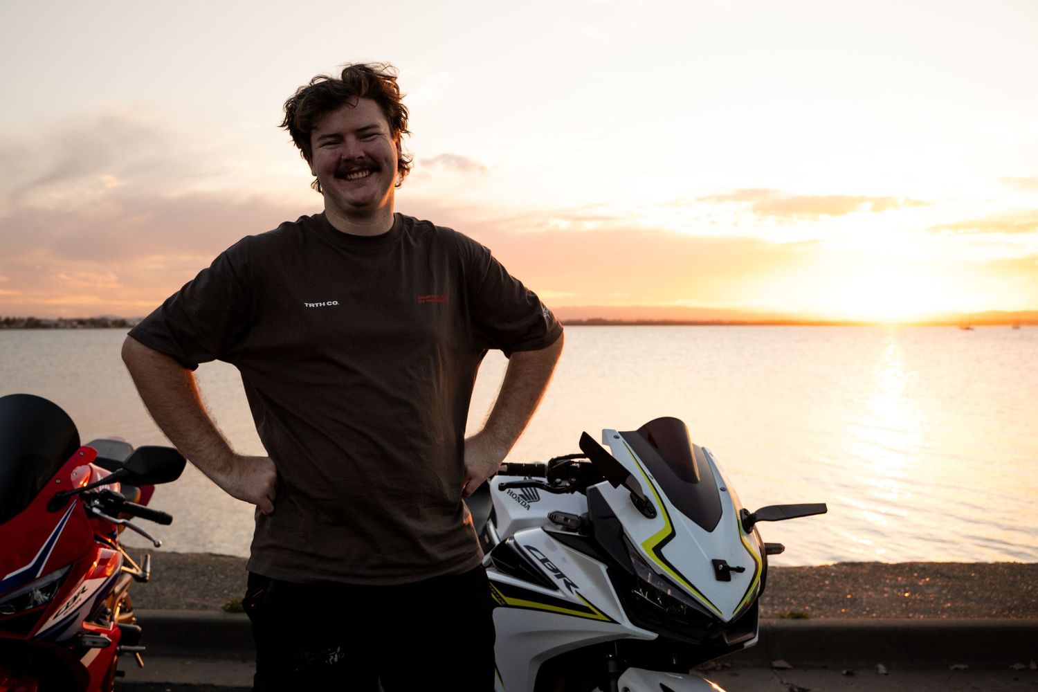 Man with mustache standing by motorcycles at waterfront during golden sunset.