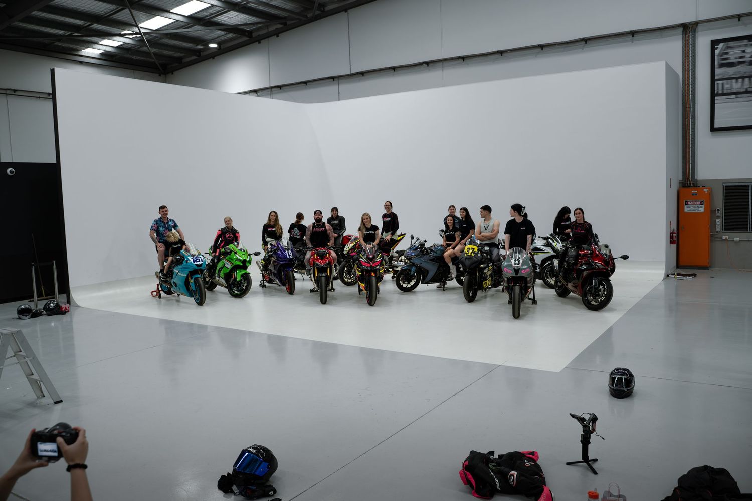 Group of motorcycle riders posing with colorful bikes in front of a white backdrop in a large studio.