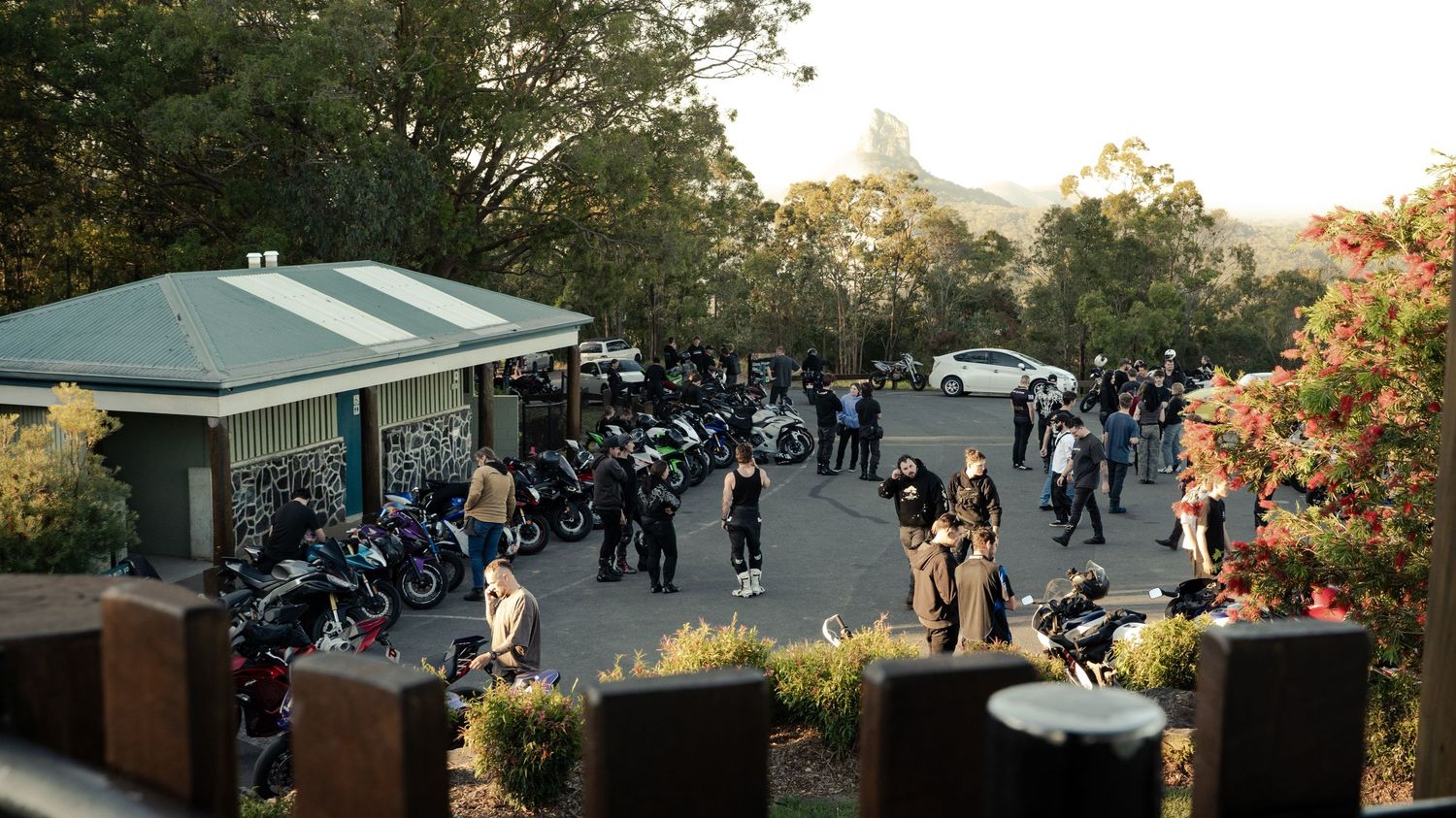 Motorcycle riders gathering in a parking lot surrounded by trees and a small building during a group ride meetup.