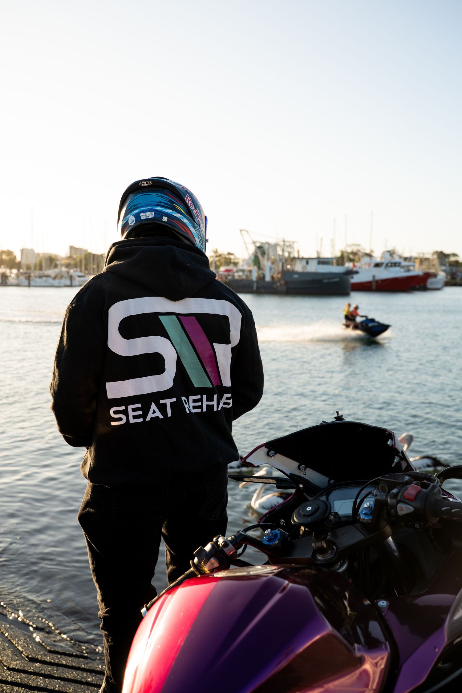 Motorcyclist in a black Seat Nehas hoodie and helmet stands by waterfront with a jet ski passing in the background.