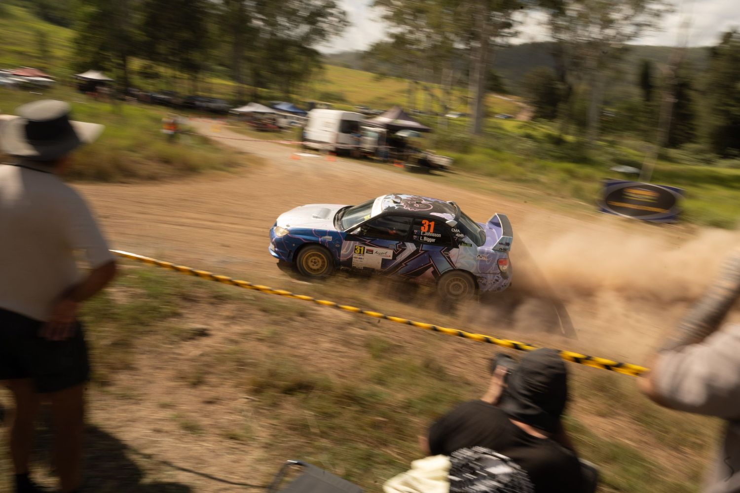 A rally car speeds through a dusty dirt track during a rally race, with spectators watching from behind safety barriers.