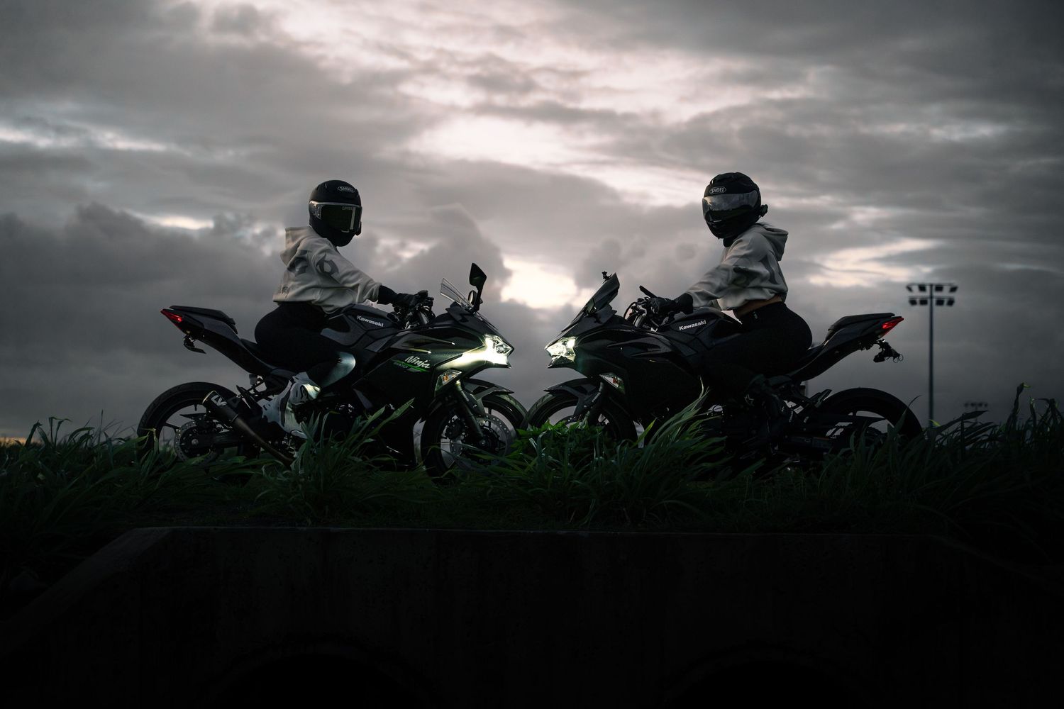 Two motorcyclists in helmets sit on sport bikes silhouetted against a dramatic cloudy sky at dusk.