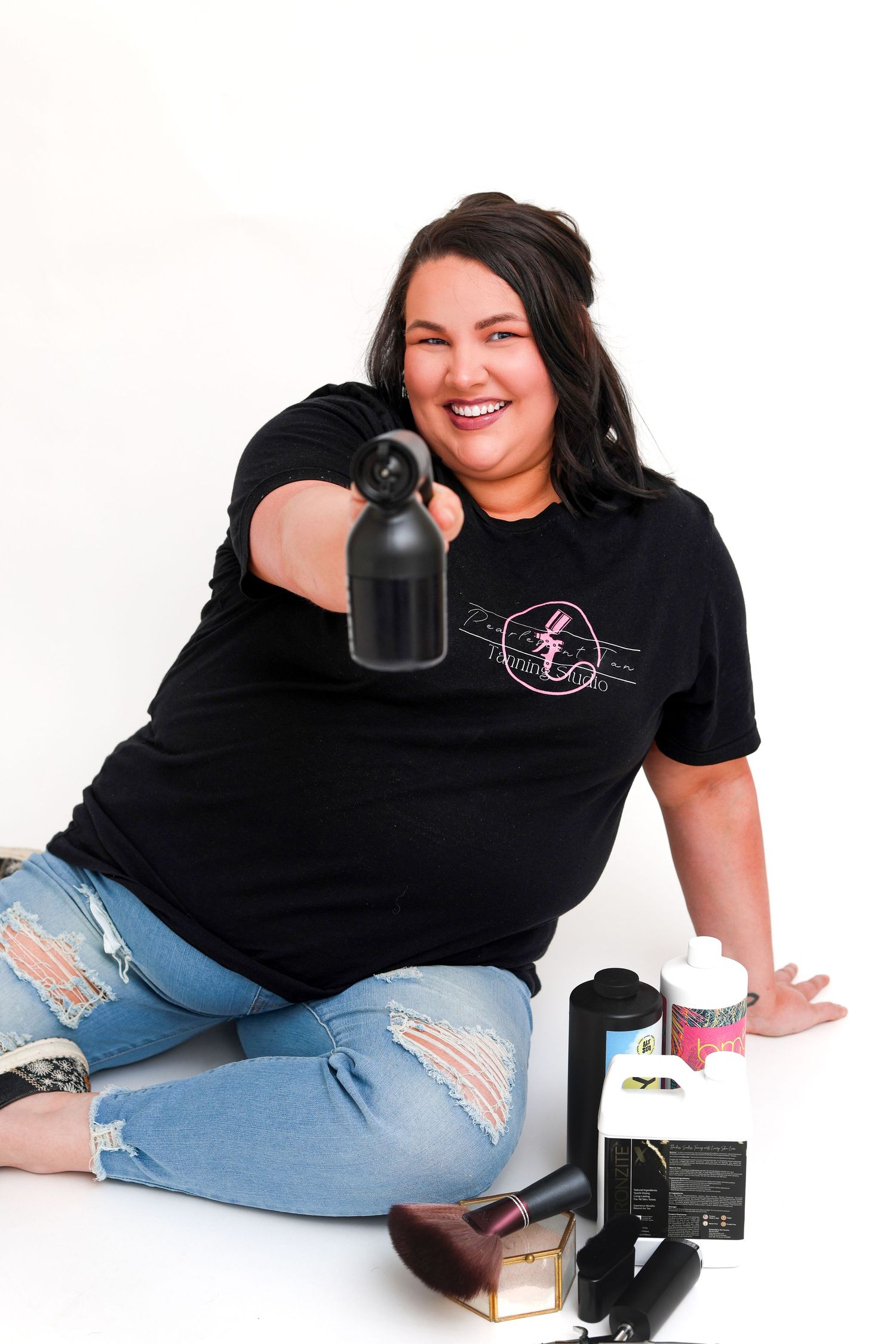 Cheerful plus-size woman in black tee sitting on floor holding beauty tool with products nearby.