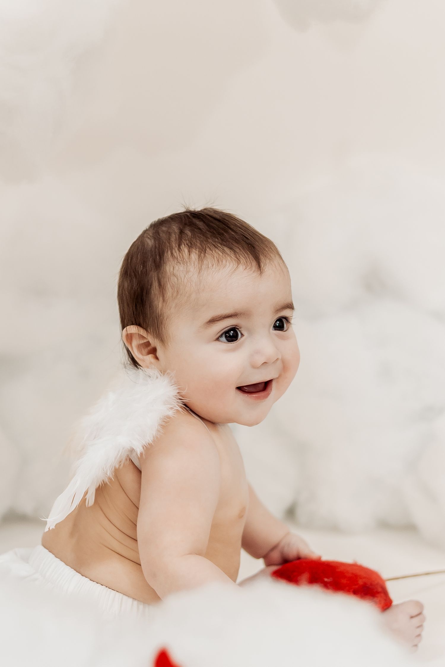 Adorable baby with white angel wings sitting and smiling, holding a red feathered arrow prop.