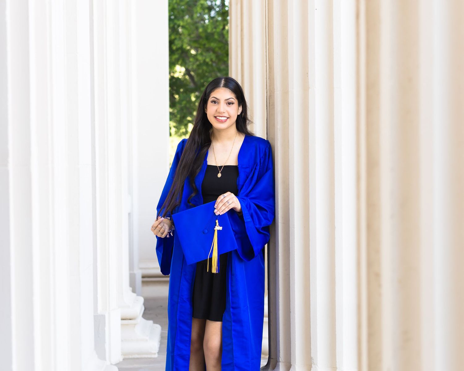 Young woman in blue graduation gown and cap smiling between white columns at graduation photo shoot.