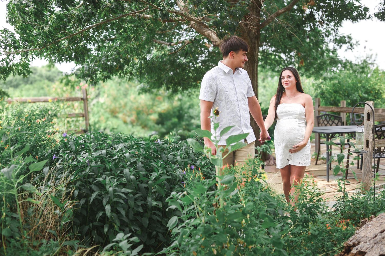 Pregnant woman in white dress walking with partner through green garden landscape.