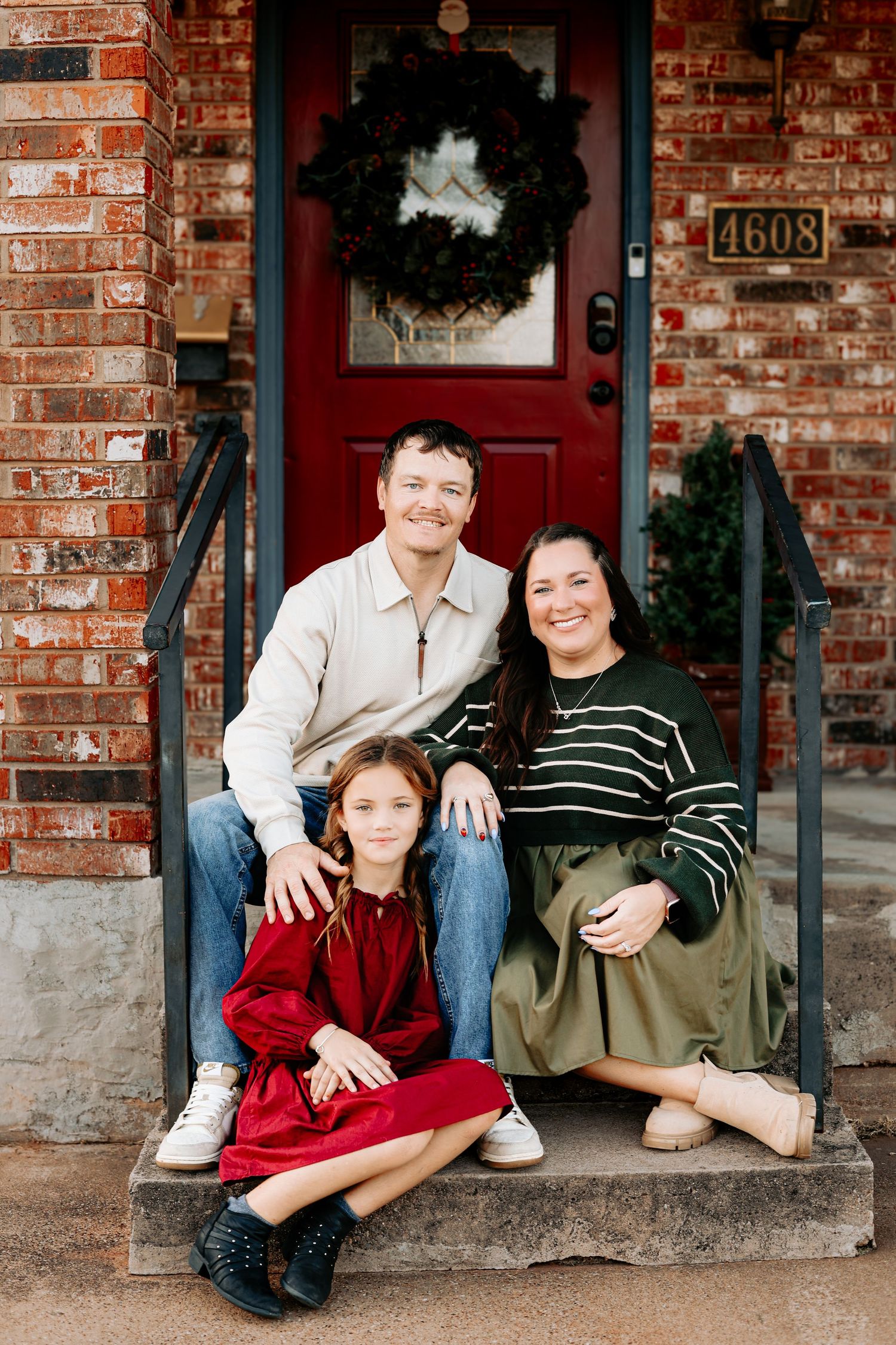 A family of three poses on brick home front steps with a red door and holiday wreath behind them.